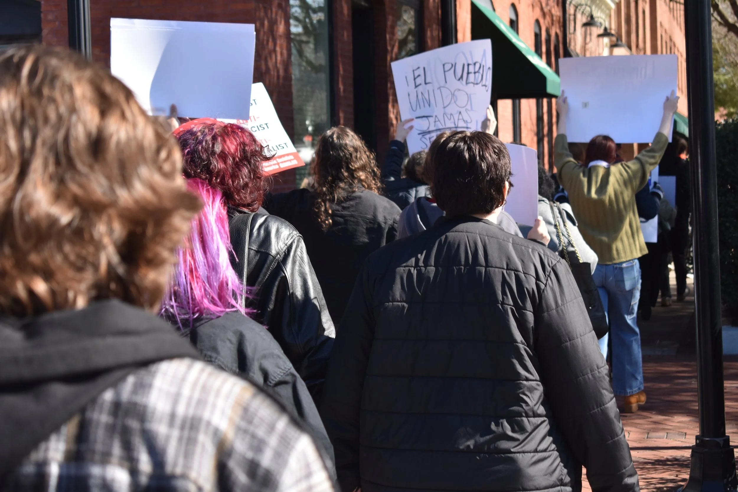 A group of people participating in a protest or march, carrying various signs. The crowd is moving in a line along a sidewalk with brick buildings and trees in the background.