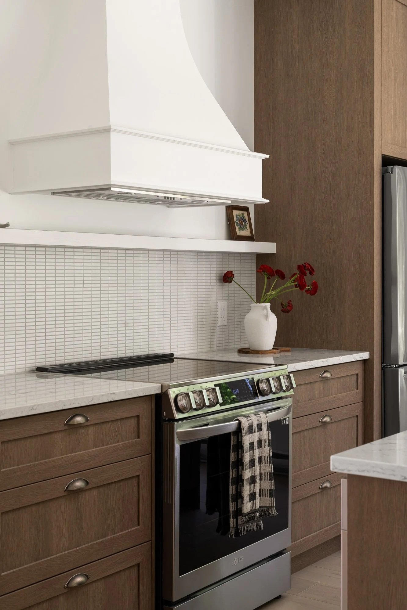 Une cuisine moderne avec des meubles en bois, une cuisinière en acier inoxydable, une hotte aspirante blanche, un plan de travail en marbre blanc, une vase blanche avec des fleurs rouges, et une petite peinture sur le mur.