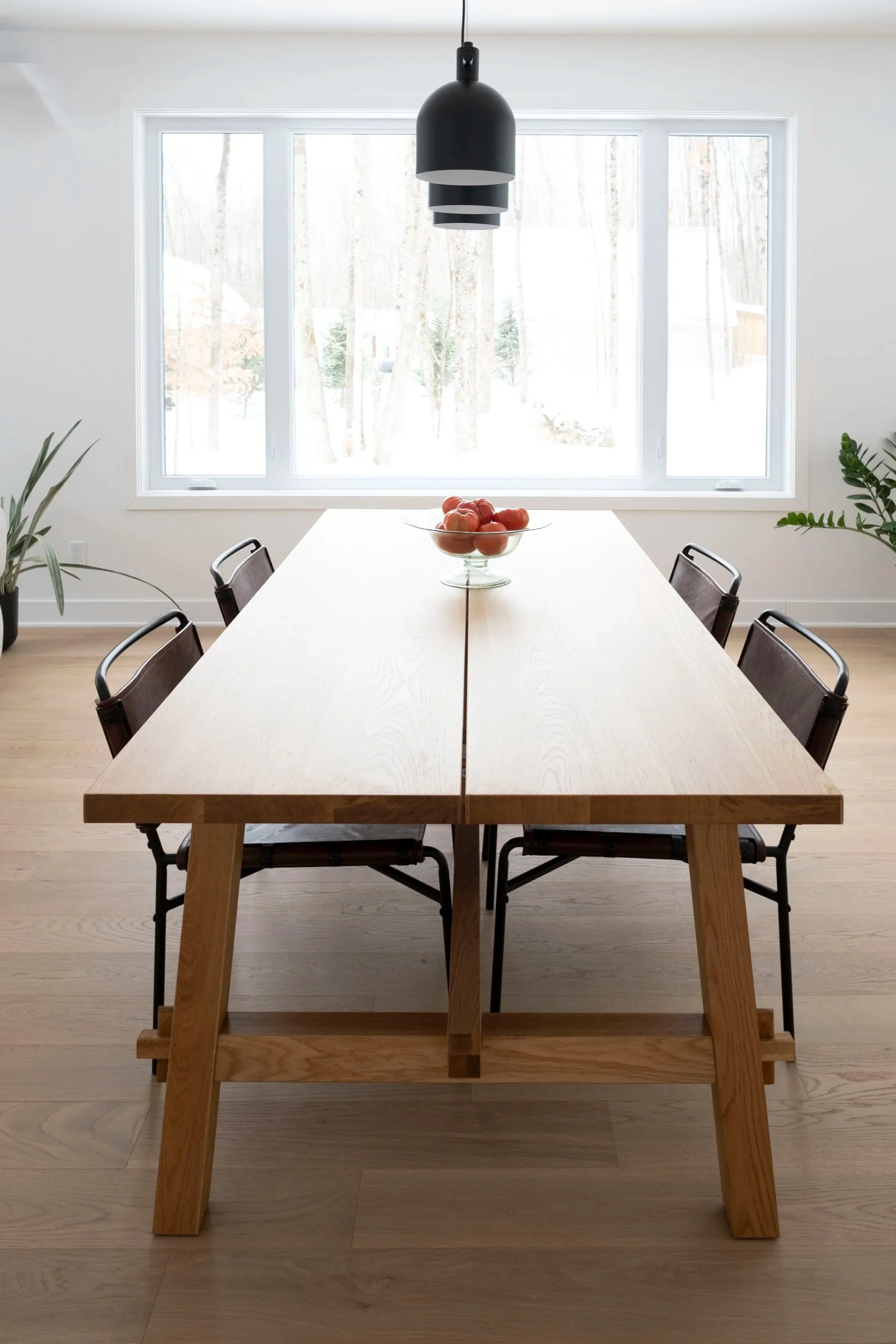 Table en bois clair avec un bol de tomates, situé dans une salle lumineuse avec grande fenêtre et plantes d'intérieur.