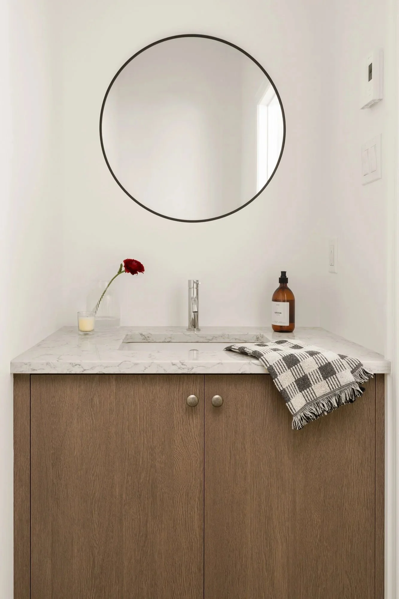 Un lavabo blanc avec comptoir en marbre, une bouteille de savon brun, une petite vase avec une fleur rouge, un miroir rond au mur, une serviette à carreaux noir et blanc sur le comptoir, murs blancs et fenêtre à lumière naturelle.