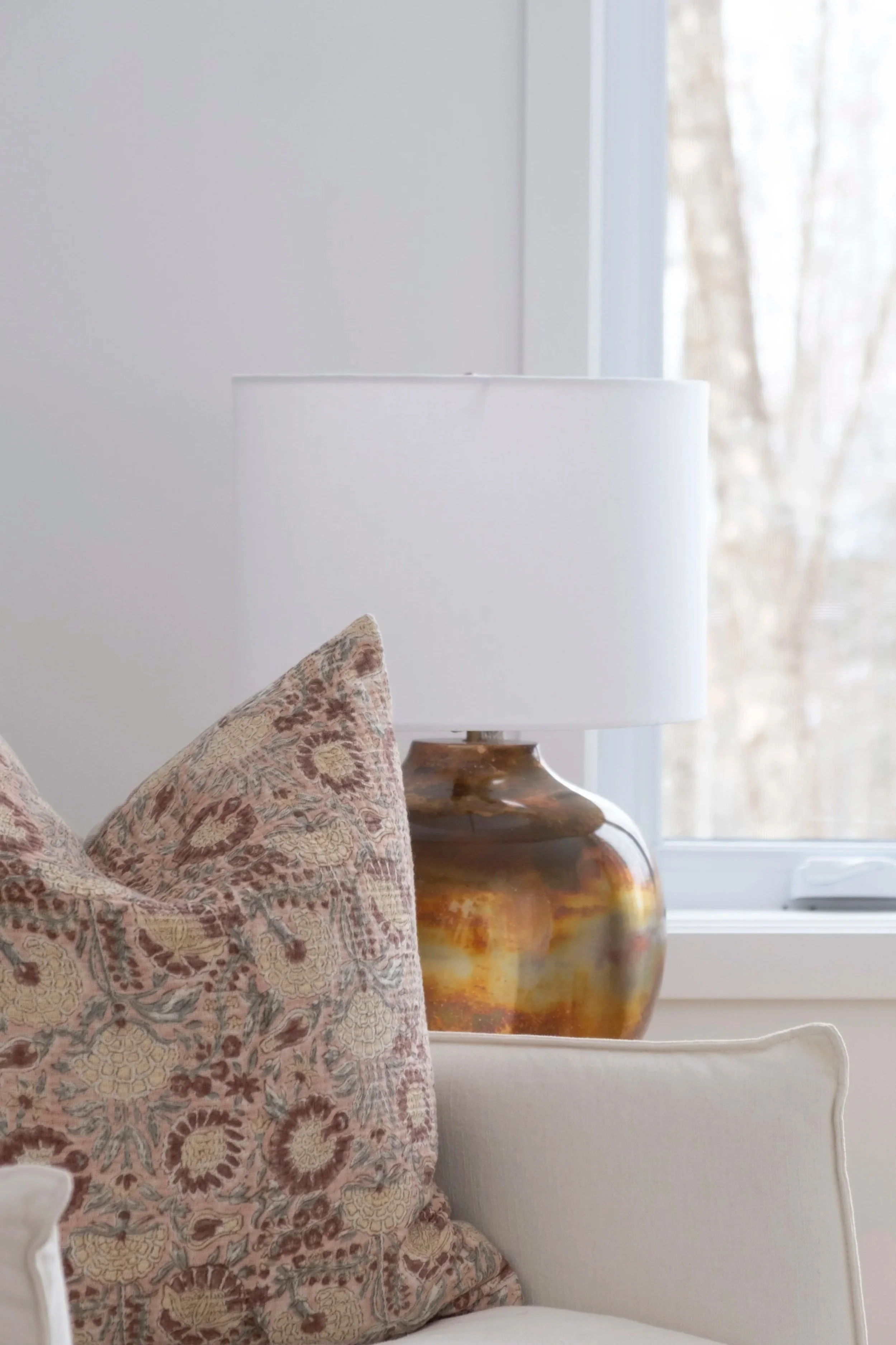 Une pièce lumineuse avec un lampadaire blanc sur une table en bois, près d'une fenêtre laissant entrer la lumière naturelle, avec un coussin à motifs floraux sur un canapé beige.