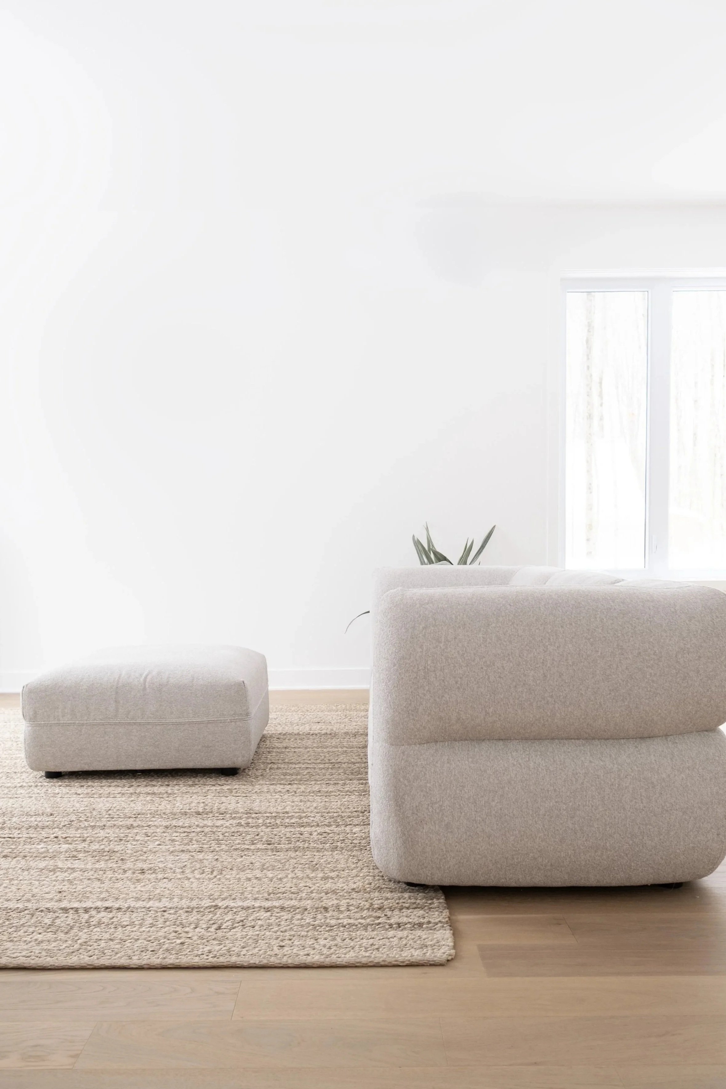 Salon minimaliste avec un fauteuil beige, un pouf assorti, un tapis tissé, et une plante verte près d'une fenêtre laissant entrer la lumière naturelle, décor épuré en blanc et beige.