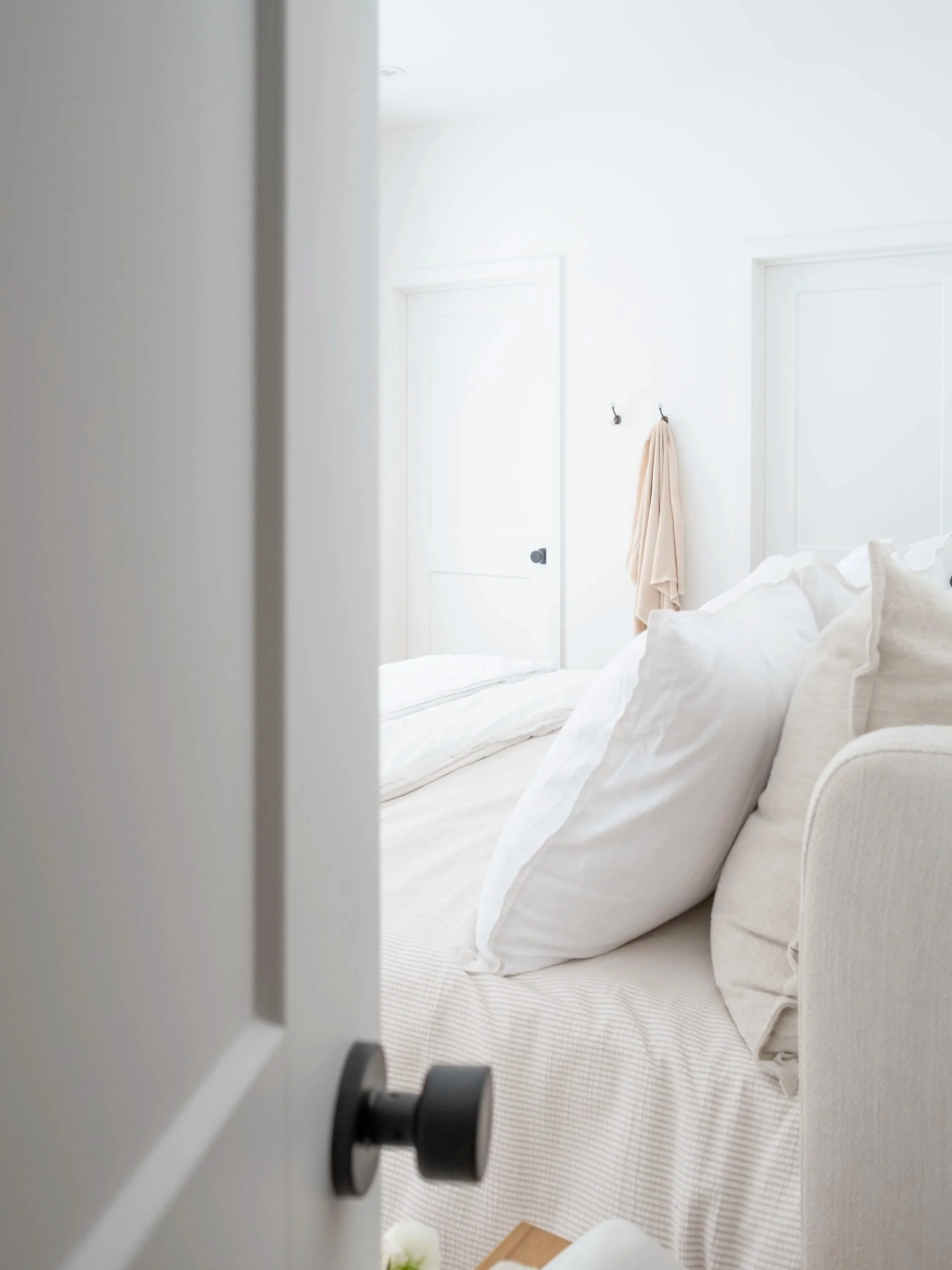 Une chambre blanche avec un lit fait, décorée de coussins et d'une couverture beige. Il y a deux crochets avec une serviette beige accrochée sur le mur. La porte blanche est entrouverte, laissant entrevoir le chiffon du lit. La scène est lumineuse, a