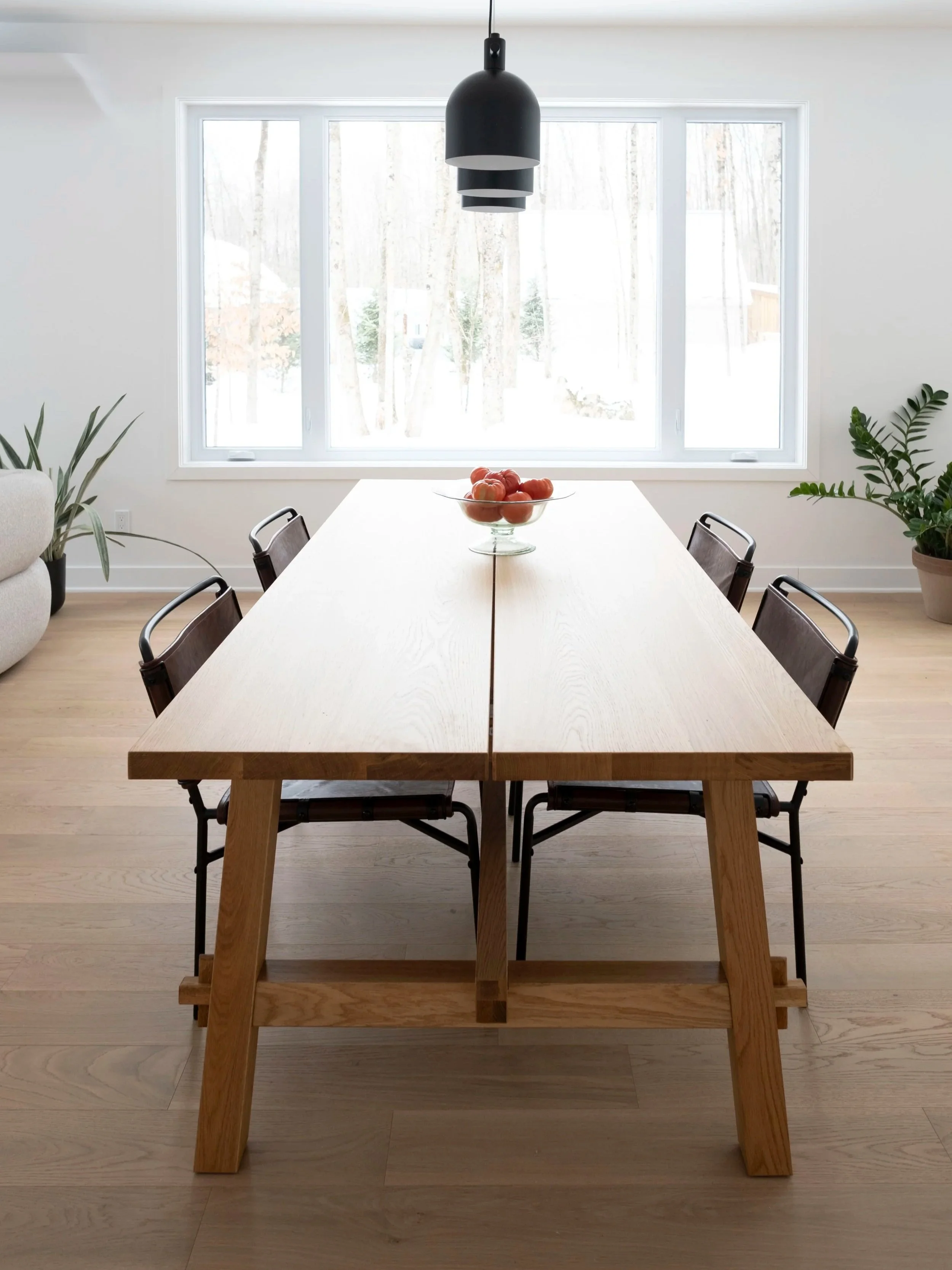 Une table en bois avec un bol de fruits rouges au centre, entourée de chaises en bois et métal. Grandes fenêtres laissant passer la lumière naturelle, végétation à l'extérieur, éclairage suspendu noir au plafond.