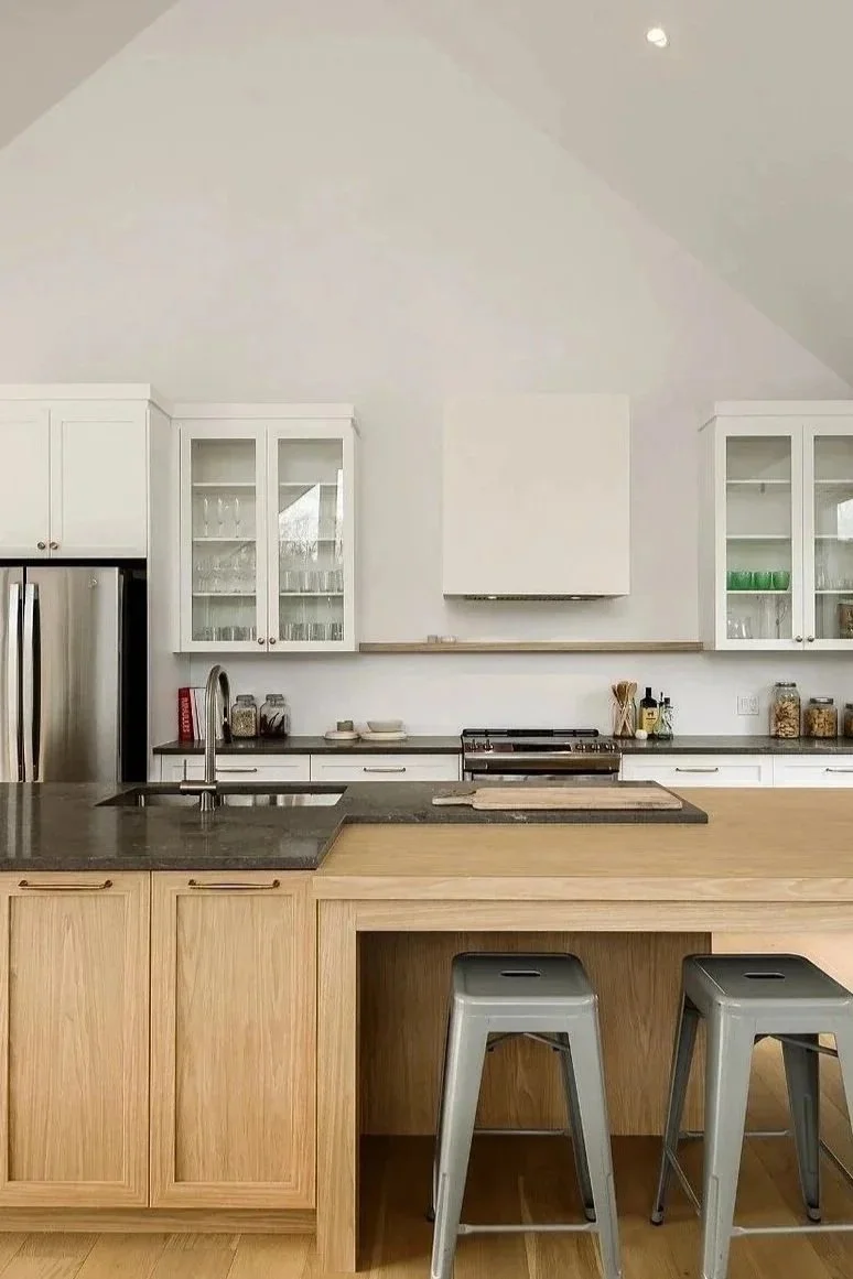 Cuisine moderne avec un îlot en bois naturel, plan de travail en pierre sombre, armoires blanches, vaisselle derrière les vitres, deux tabourets en métal gris et un mur blanc avec une hotte encastrée.
