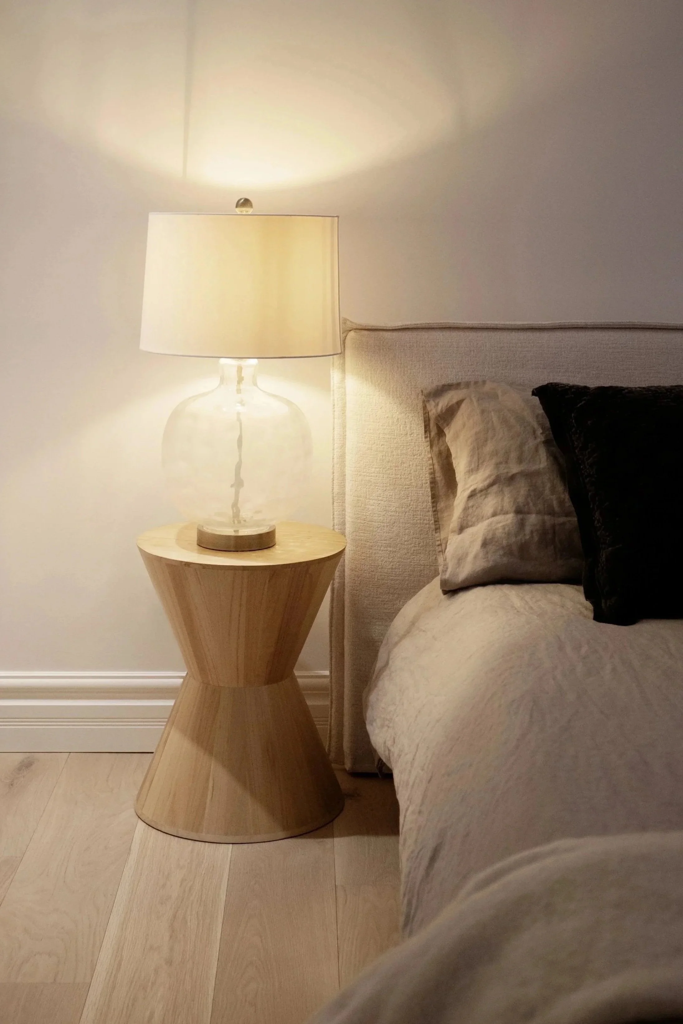 Une lampe de table en verre avec un abat-jour beige sur une petite table en bois à côté d'un lit avec des coussins beige et noirs dans une chambre.