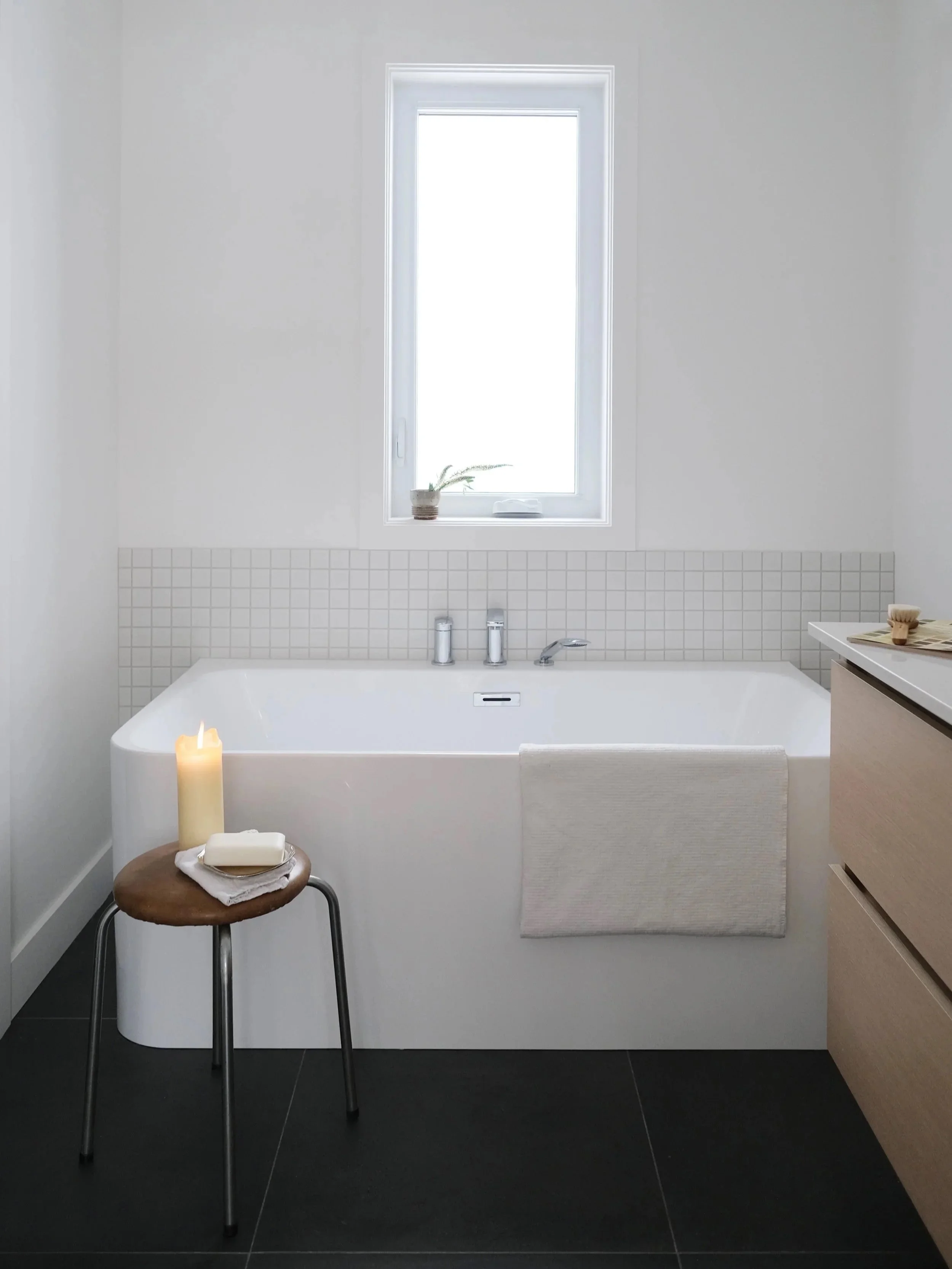 Salle de bain minimaliste avec une baignoire blanche, une fenêtre, une petite plante en pot sur le rebord de la fenêtre, une bougie allumée sur un tabouret en bois, un tapis blanc suspendu à la baignoire, et des meubles en bois clair.