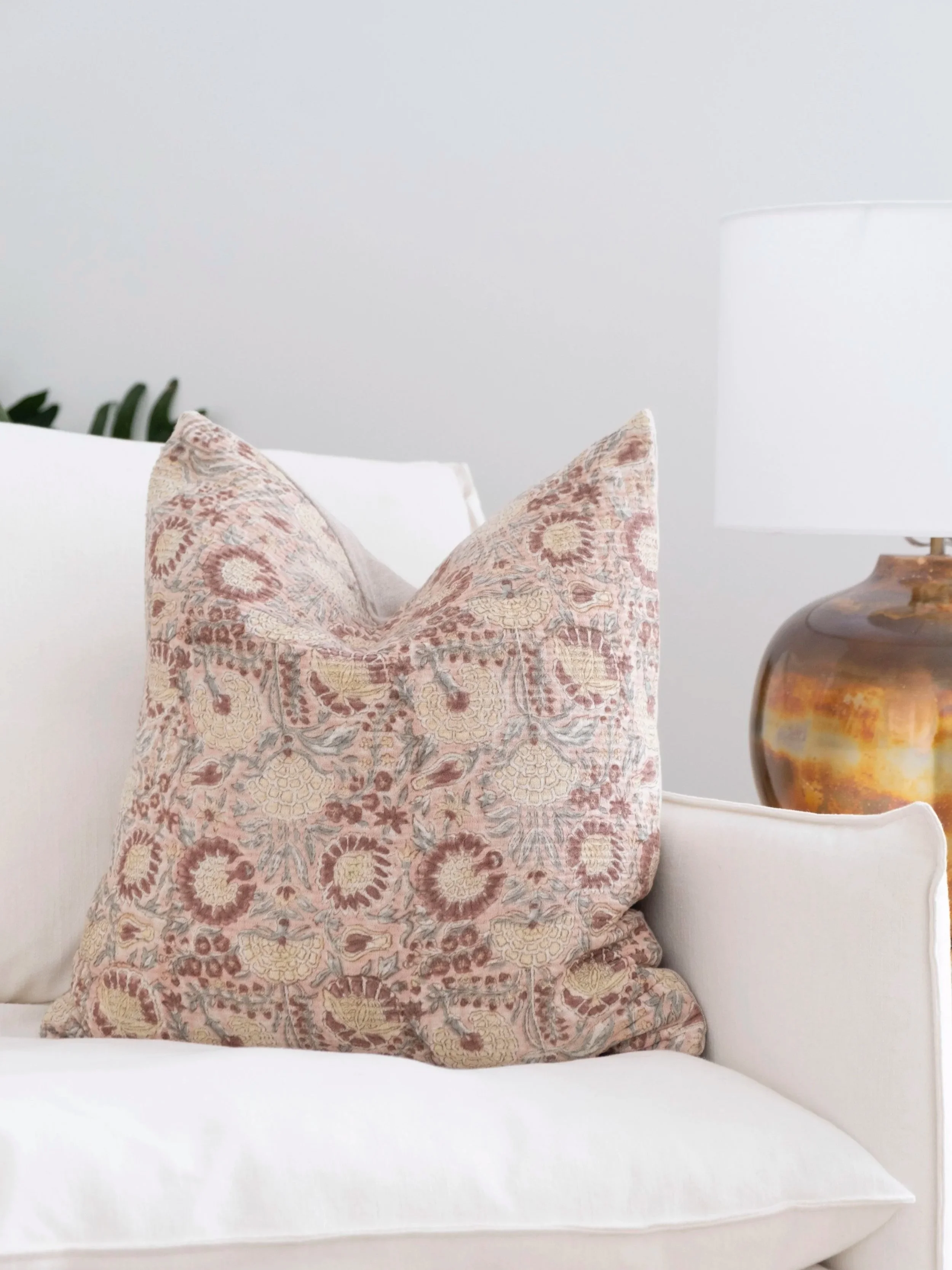 Coussin aux motifs floraux pastel posé sur un canapé blanc, avec une lampe en céramique à côté et un mur gris clair en arrière-plan.