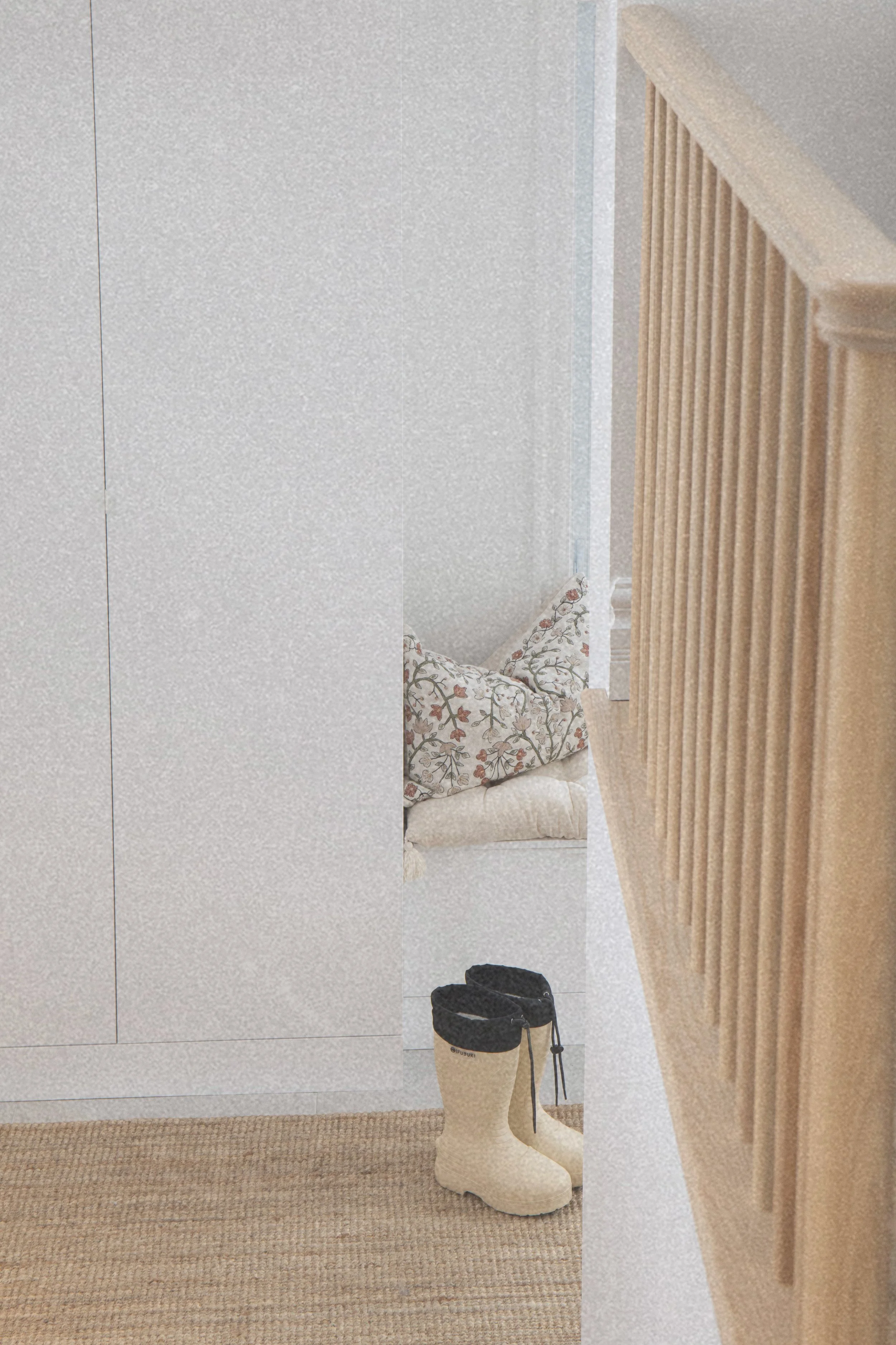 Une vue rapprochée d'une entrée de maison épurée, avec une rampe d'escalier en bois clair à lattes et des armoires de rangement sur mesure. Un tapis de jute et une apire de bottes de pluie beiges créent un coin fonctionnel et accueillant.