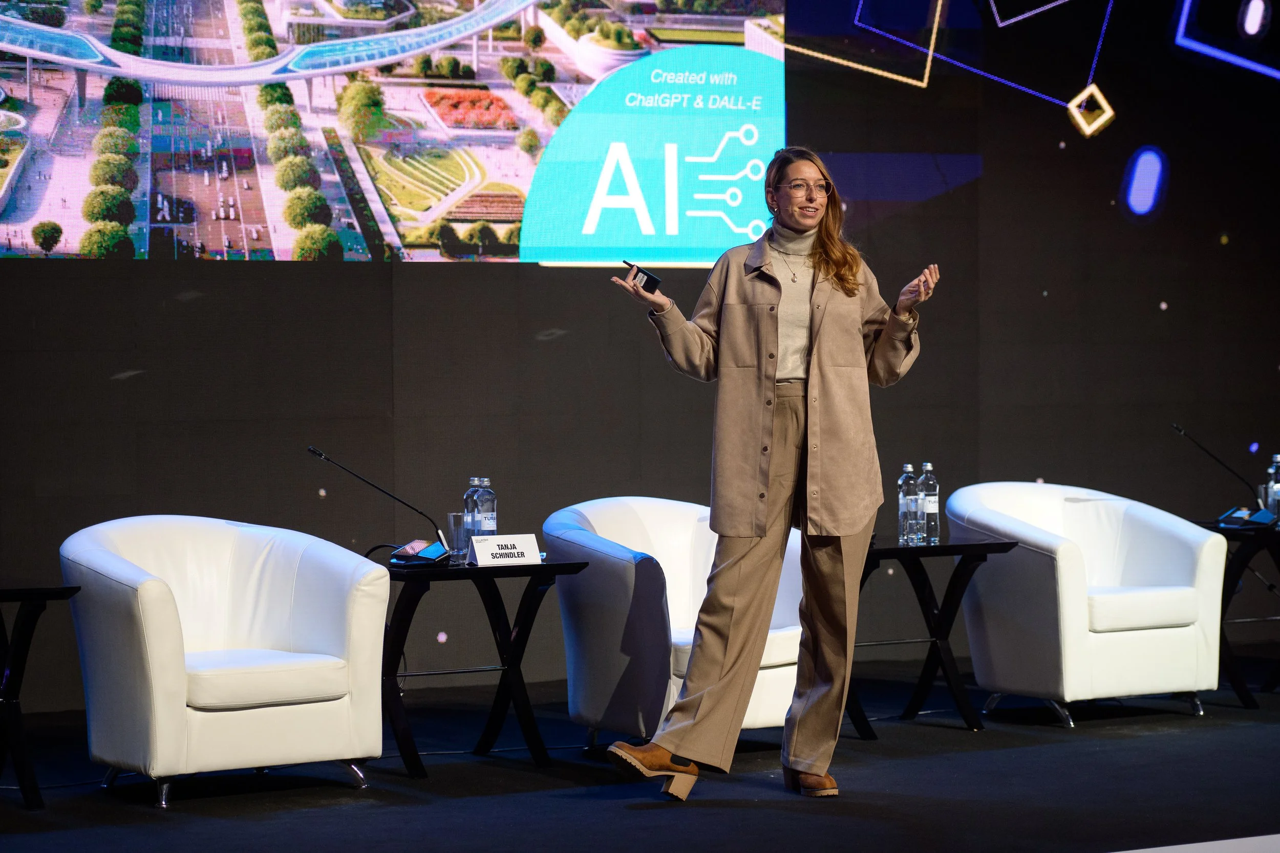 Junge Frau spricht auf einer Bühne während einer Konferenz über KI, mit einem großen Bildschirm im Hintergrund, auf dem das Wort 'AI' und technologische Grafiken zu sehen sind, umgeben von leeren weißen Sessel und Tischen mit Wasserflaschen.