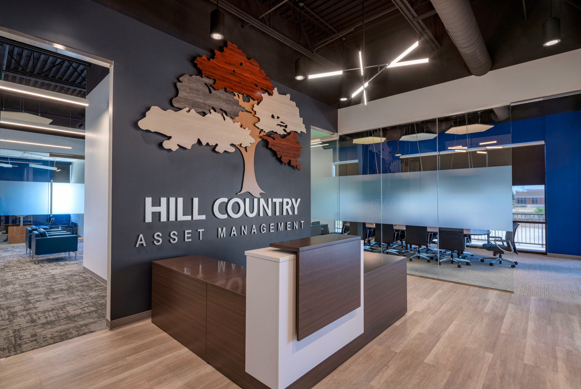 Modern office lobby with a wall display featuring a cutout map of a region in wood, labeled 'HILL COUNTRY ASSET MANAGEMENT.' The office has glass walls, a seating area with blue chairs, and contemporary lighting fixtures.