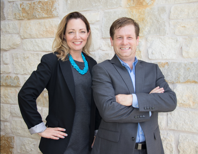 ResolveCos. LLC Managing Principals, Russell R Collins and Danielle H Collins. A smiling woman in a black blazer and gray dress with a blue necklace standing next to a smiling man in a gray suit with crossed arms, in front of a stone wall.