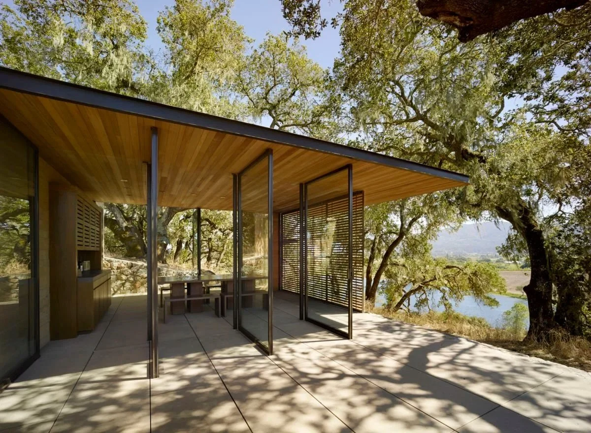 Modern house with a wooden ceiling and glass walls, surrounded by trees and overlooking a river in a natural setting.