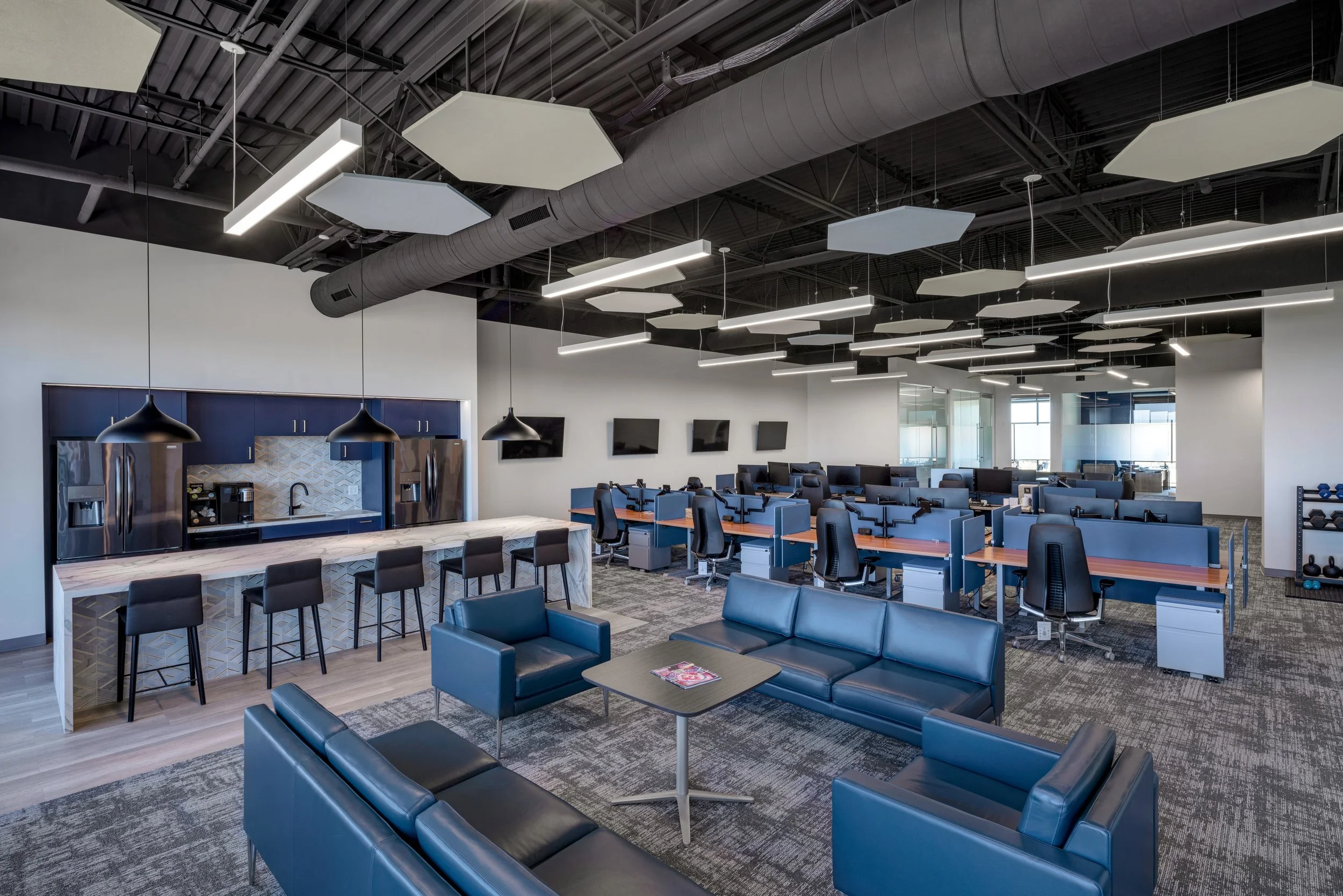 Modern office space with open layout, featuring blue seating, high desks with chairs, multiple wall-mounted monitors, kitchen area with high chairs, and unique ceiling lights.