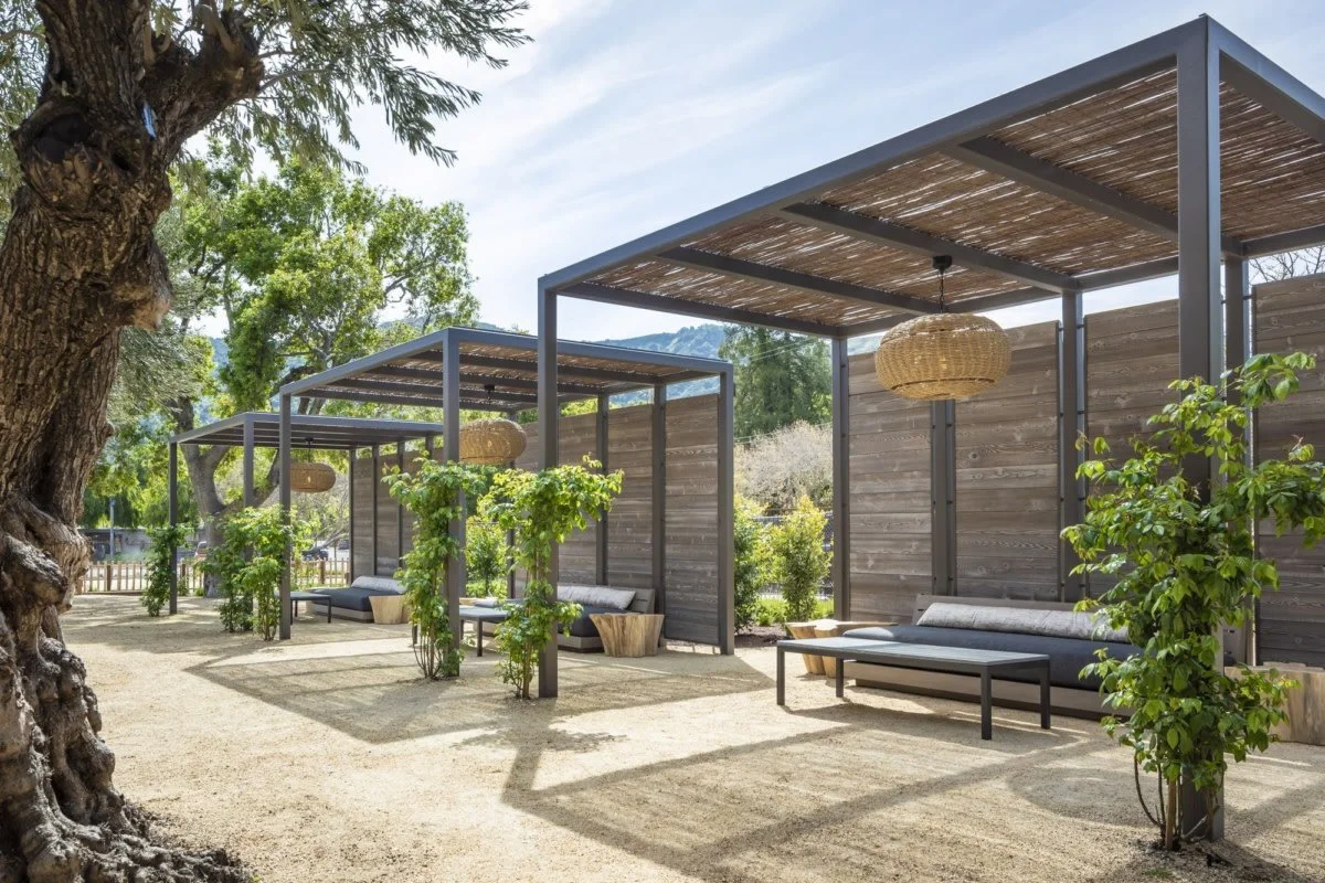 Outdoor seating area with shaded wooden structures, greenery, and hanging wicker light fixtures, set on a sandy ground with trees and a fence in the background.