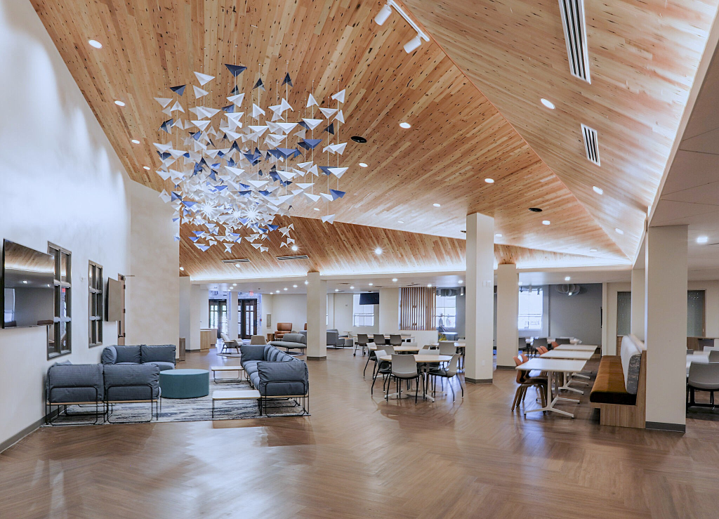 A spacious, modern common area or lounge with wooden ceiling and floor, featuring blue and gray sofas, dining tables with white chairs, and decorative hanging art made of white and blue geometric shapes.