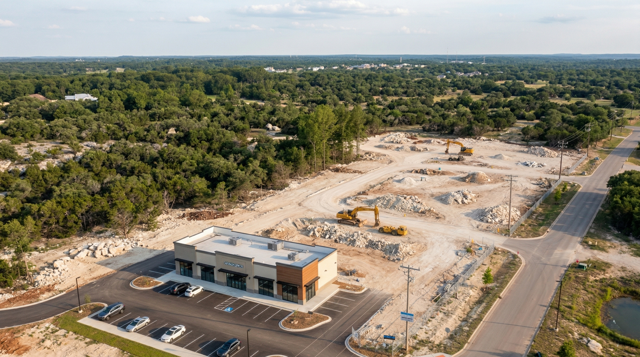 Aerial view of a construction site next to a small shopping center and surrounded by trees. Construction equipment and piles of dirt are visible, and a new building is under development.