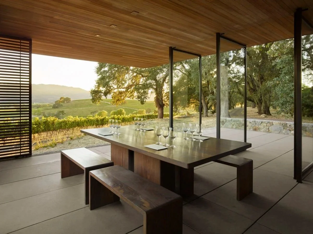 Modern dining area with large wooden table set with wine glasses and napkins, open to scenic vineyard and trees at sunset