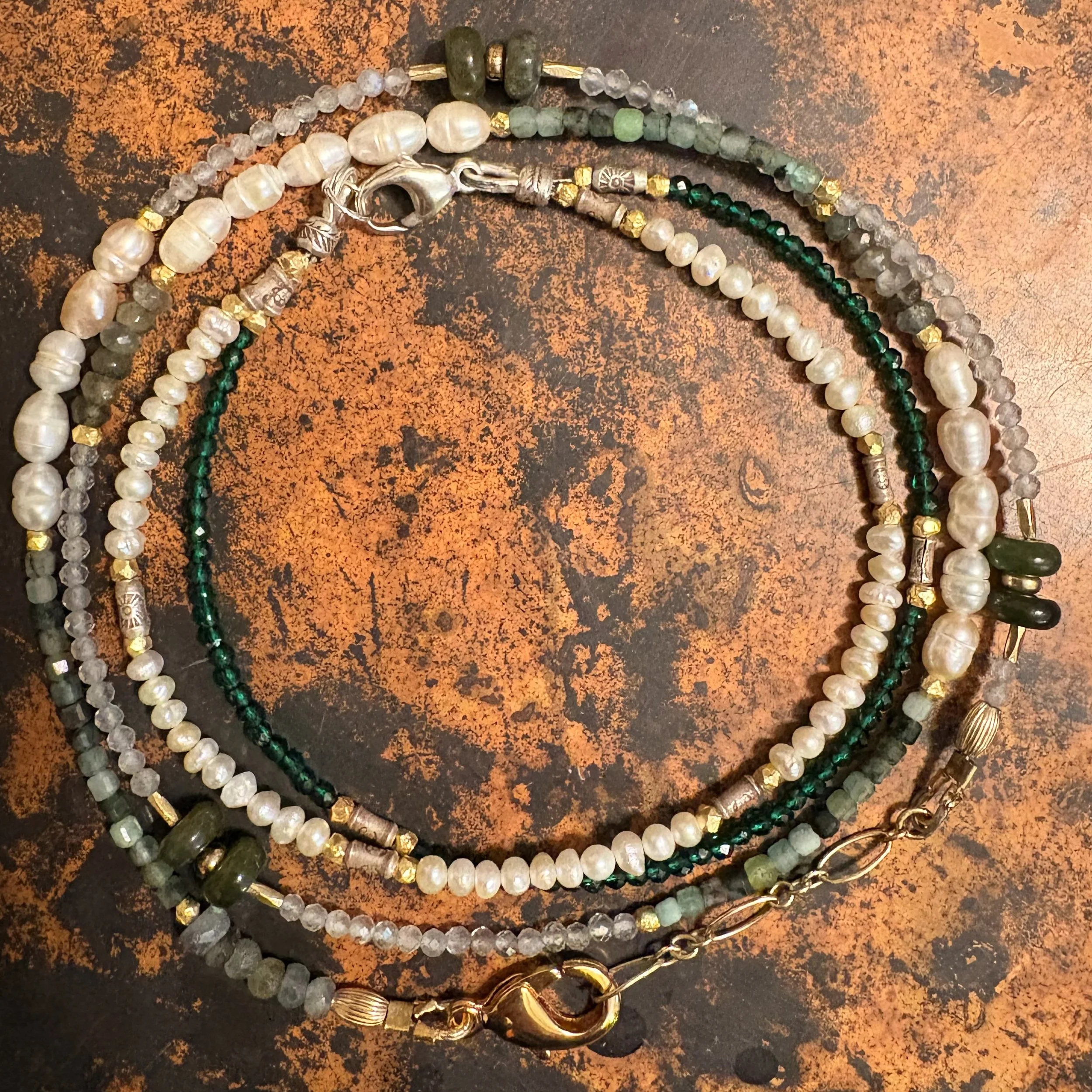 Three beaded necklaces, one with green and gold beads, one with white pearls, and one with small clear and gray beads, on a rust-colored textured surface.