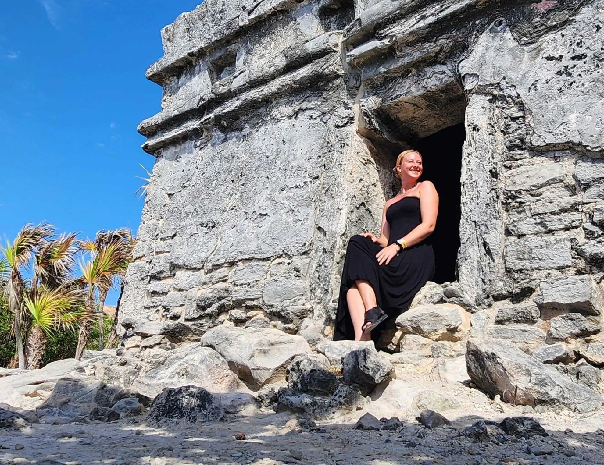A woman in a black dress sitting on rocks in front of an ancient stone structure with a woman smiling inside the window opening, against a clear blue sky and some palm trees in the background.