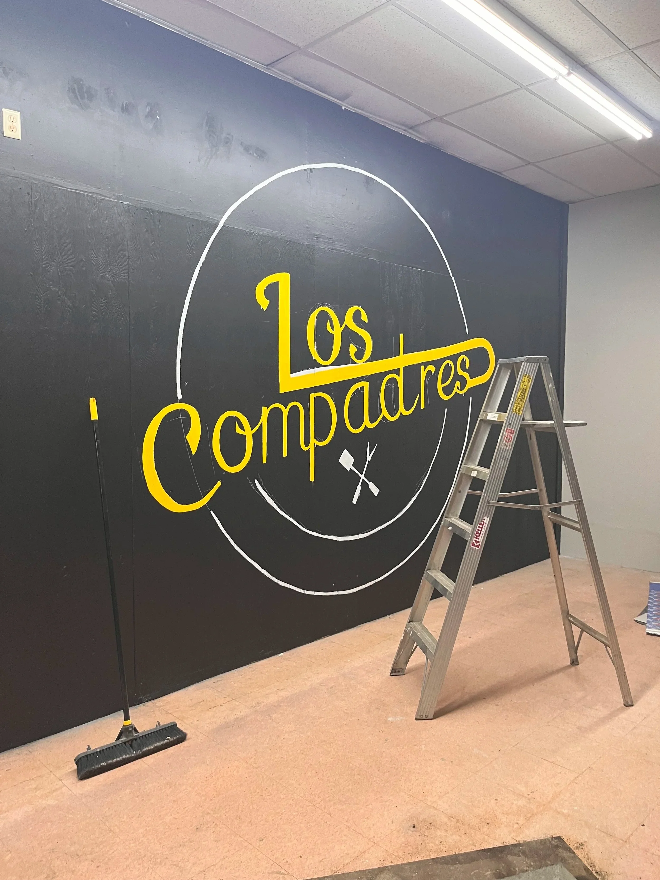 A black wall with a yellow and white hand-painted sign that reads 'Los Compartes' with a circle and arrow designs, a ladder, and a broom leaning against the wall.