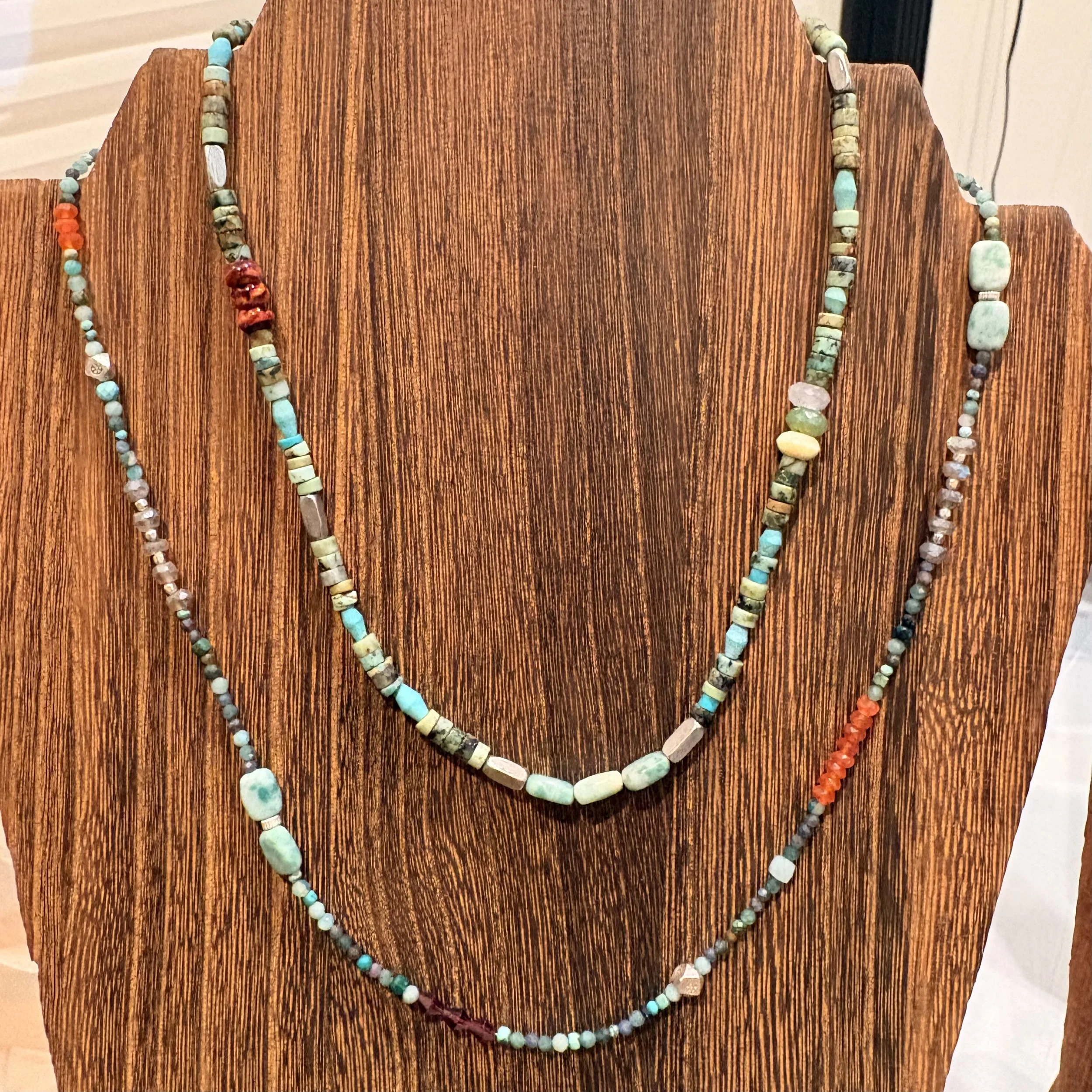 Two layered necklaces made of turquoise, coral, and other beads displayed on a wooden jewelry stand.