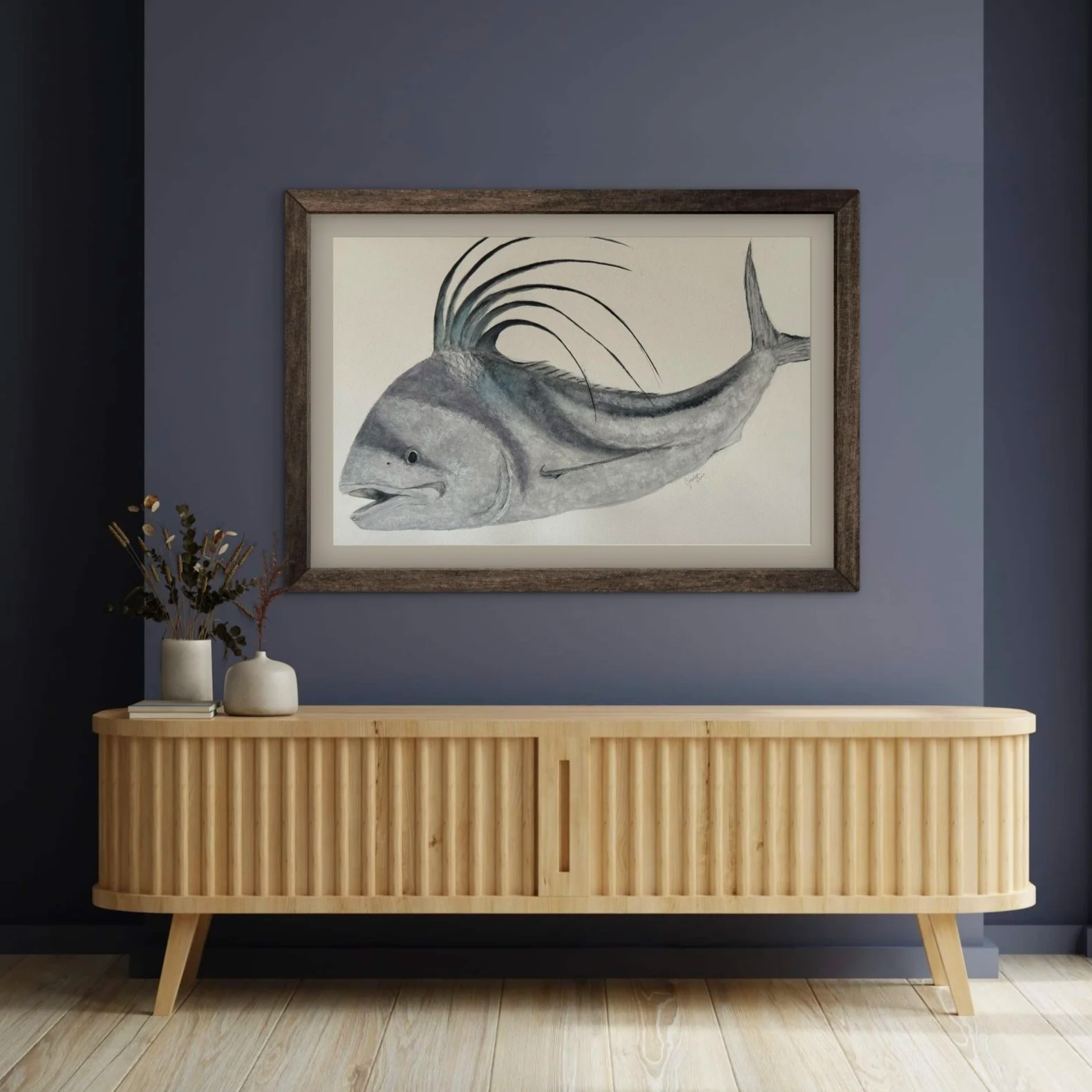A framed watercolor painting of a fish hung on a gray wall above a wooden sideboard. The sideboard has a rattan-like design on the front and legs, and displays three white vases with dried plants on the left.