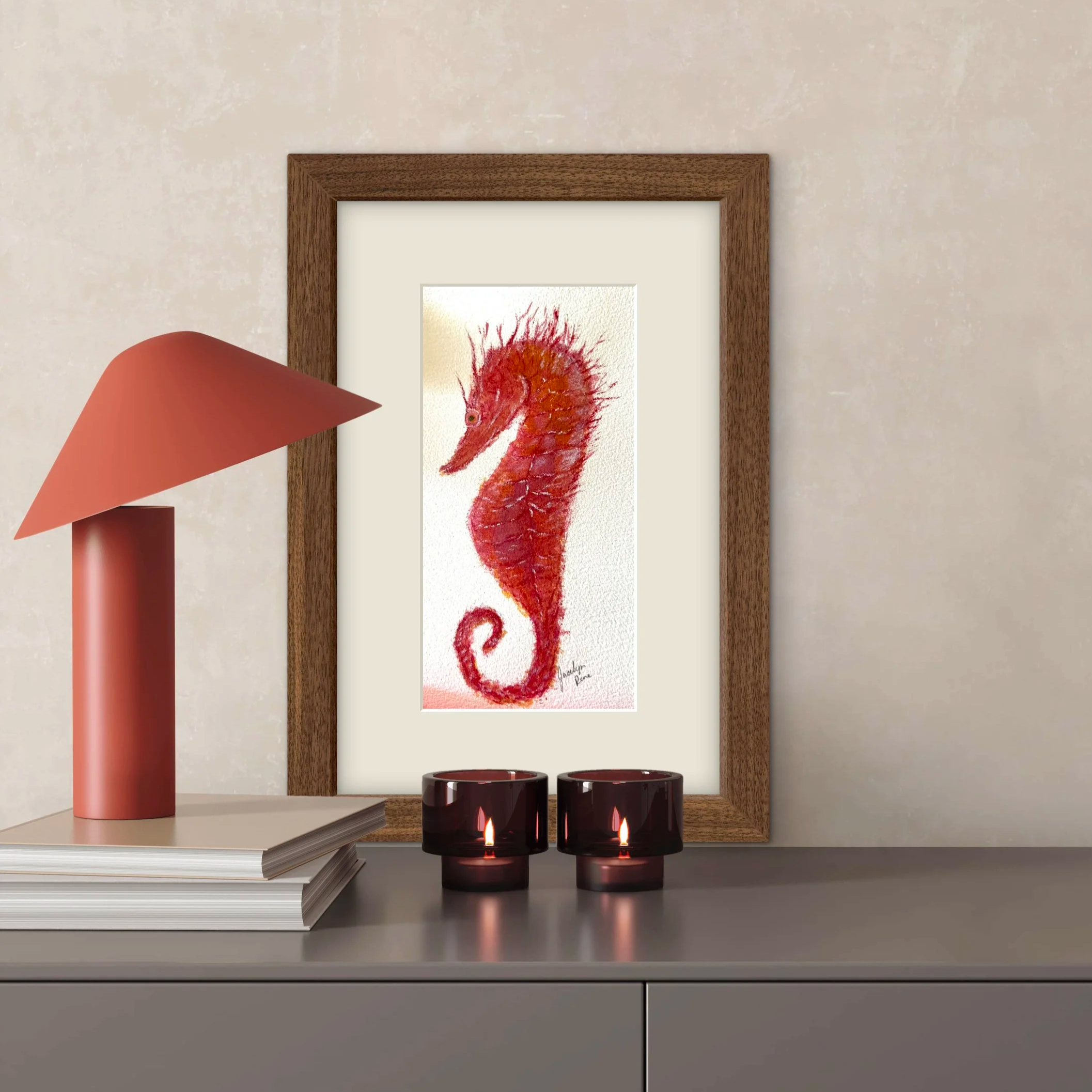Decorative tabletop arrangement featuring a framed artwork of a seahorse with a red and orange color scheme, three lit candles, a pink vase, and a stack of notebooks, all on a gray surface against a light-colored wall.