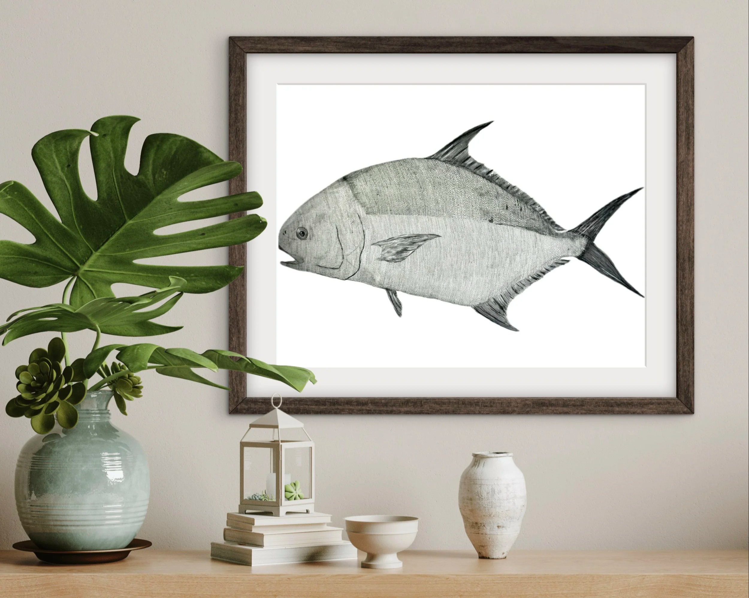 A modern interior decor scene featuring a framed black-and-white fish illustration on the wall, a large green monstera plant in a white pottery vase, a white lantern, a white decorative statue, a stack of books, and various white ceramic bowls on a w