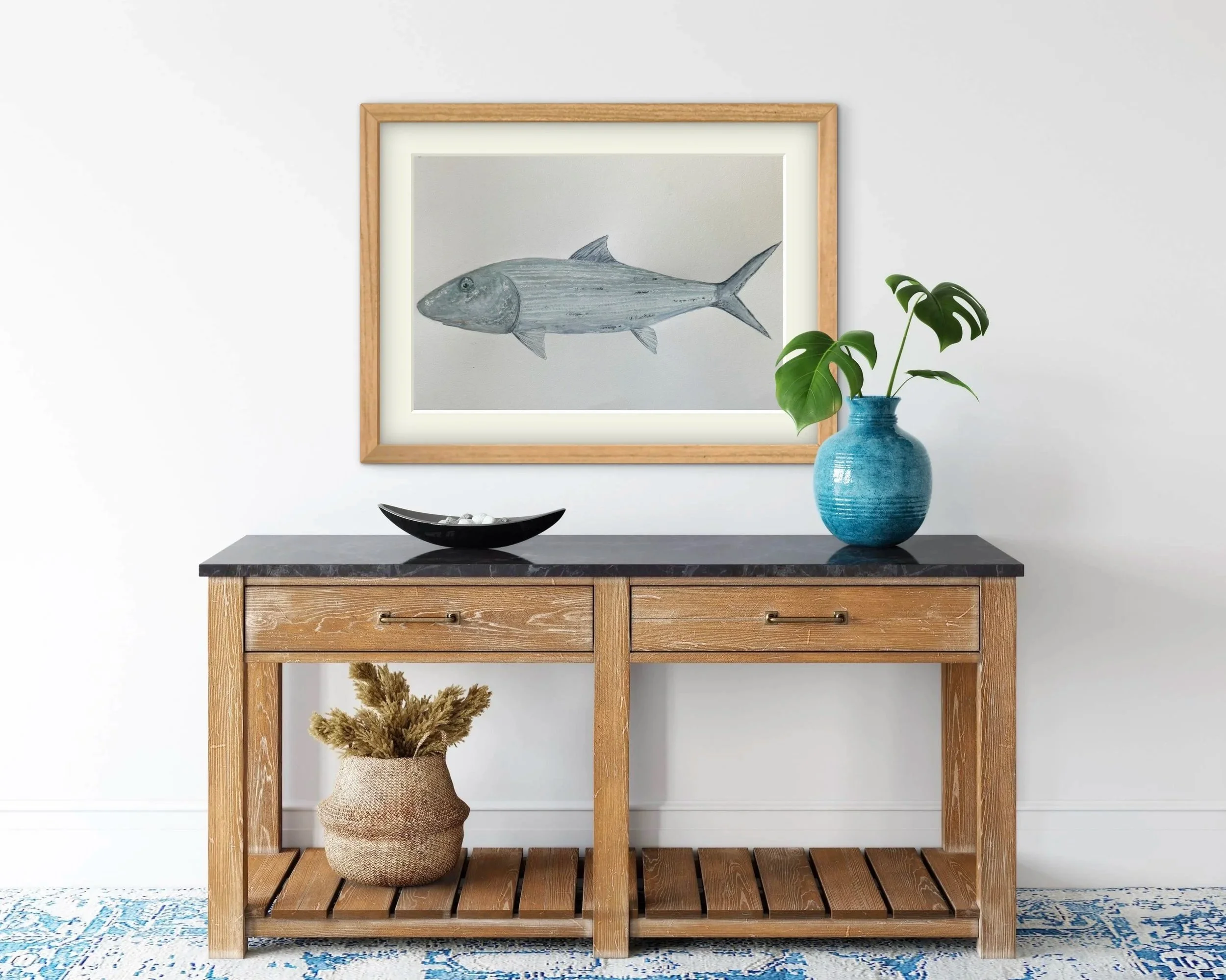 A wooden console table with black hardware, topped with a black bowl, positioned in front of a white wall. On the wall, there's a framed picture of a fish, and a blue vase holding green monstera leaves is placed on the right side of the table. A wove