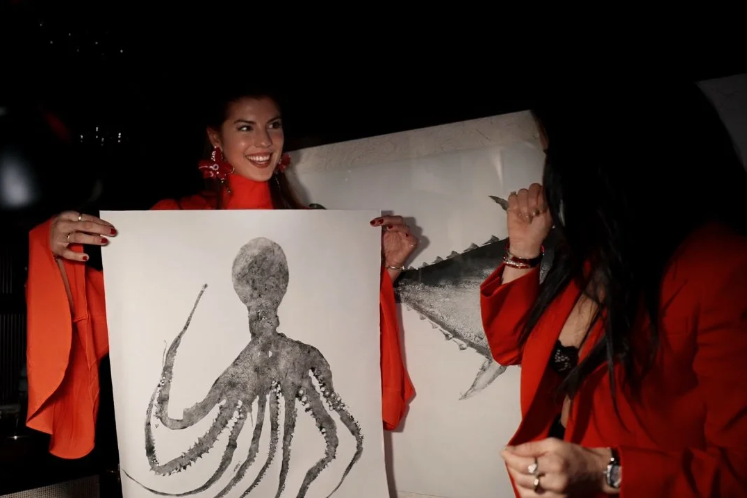 Two women wearing red are in a car at night, one holding an artwork of an octopus and the other pointing at a part of a different artwork with a whale fin.
