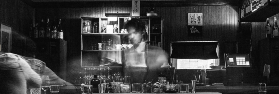 A person working behind the bar in a dimly lit restaurant, with shelves of bottles and glasses in the background.