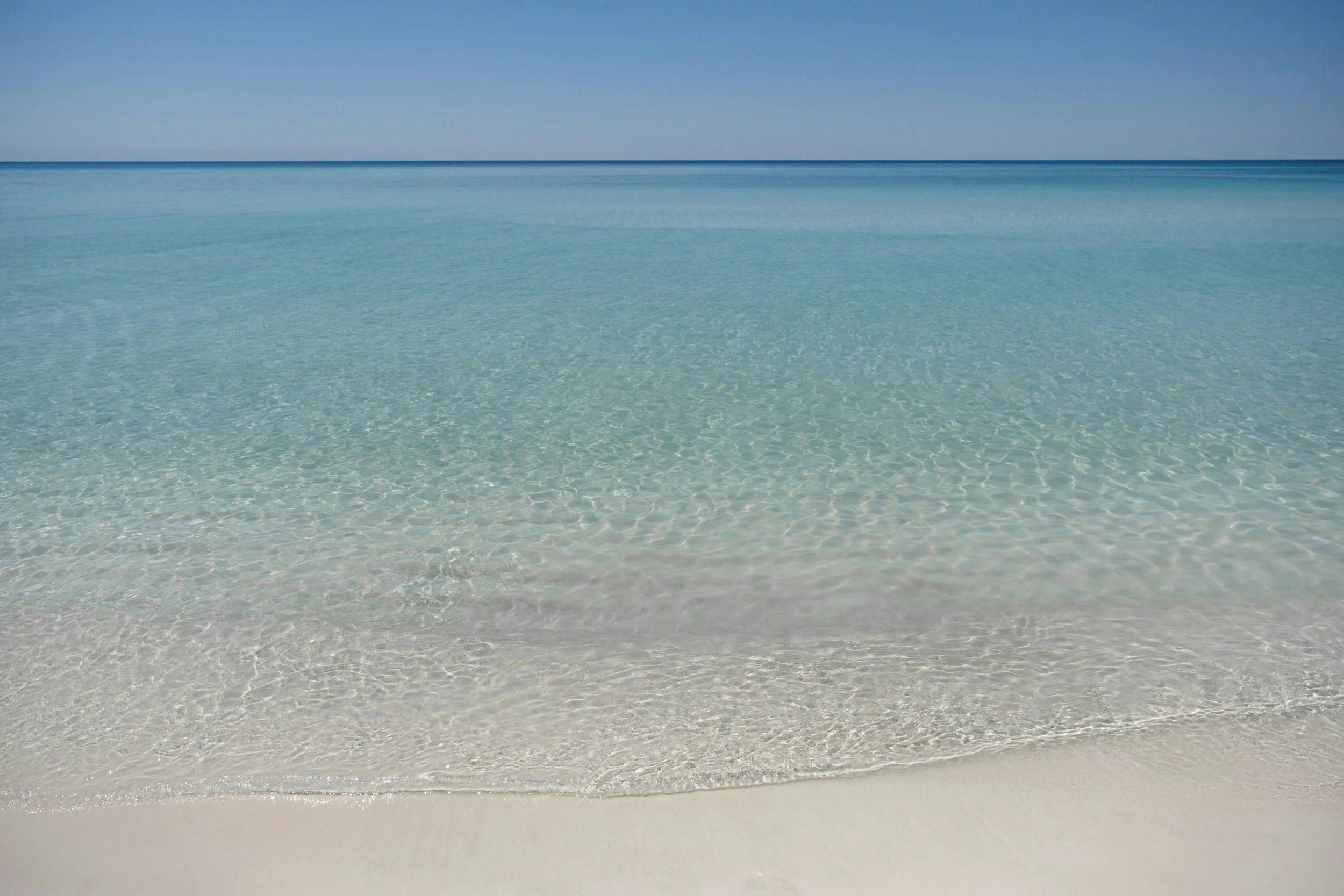 Calm turquoise ocean water lapping onto white sandy beach under clear blue sky.