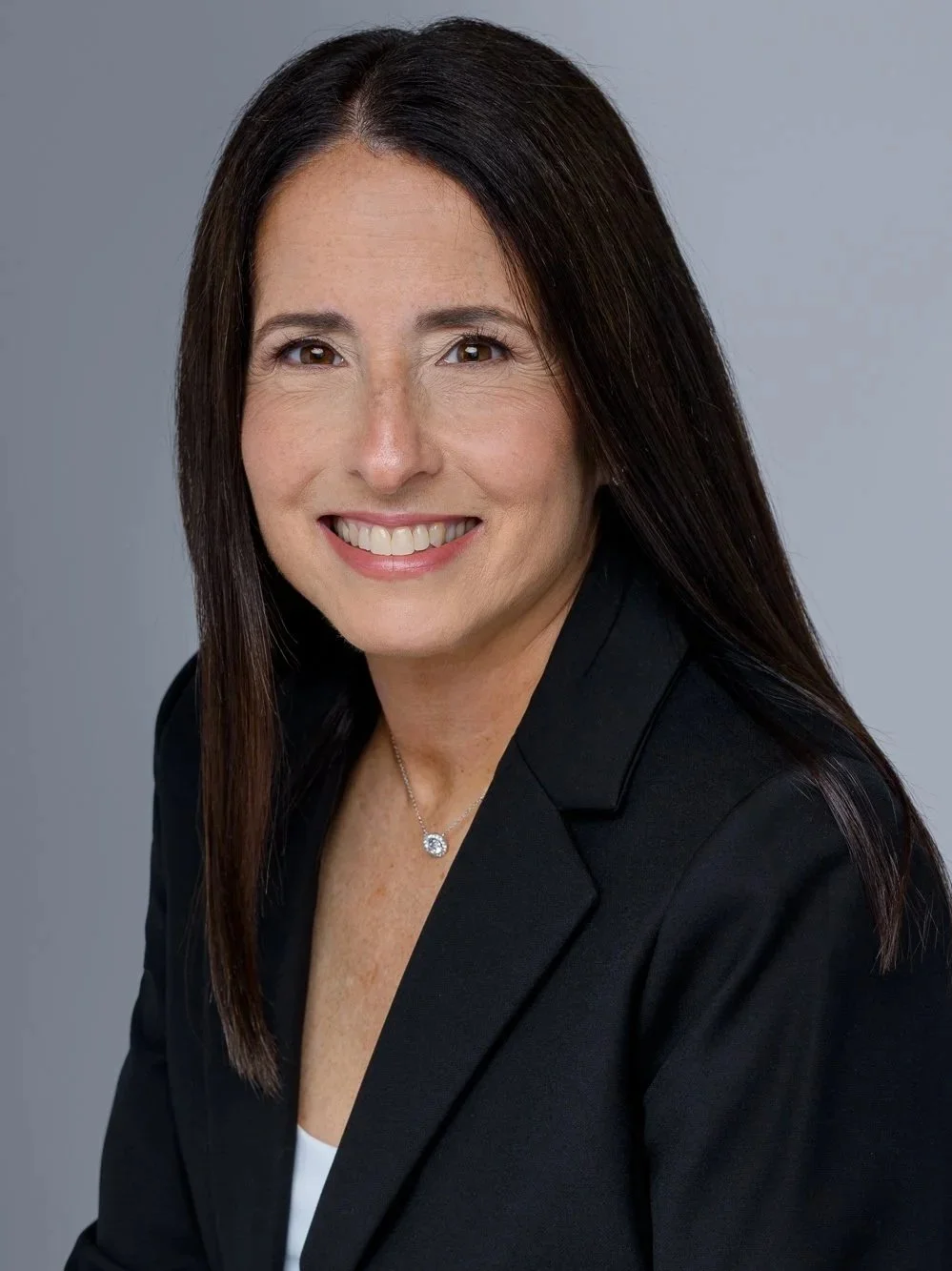 Professional woman with long dark hair, smiling, wearing a black blazer and a necklace, against a gray background.