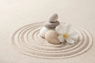 Stacked smooth gray and white stones with a white flower on sand with concentric circle patterns