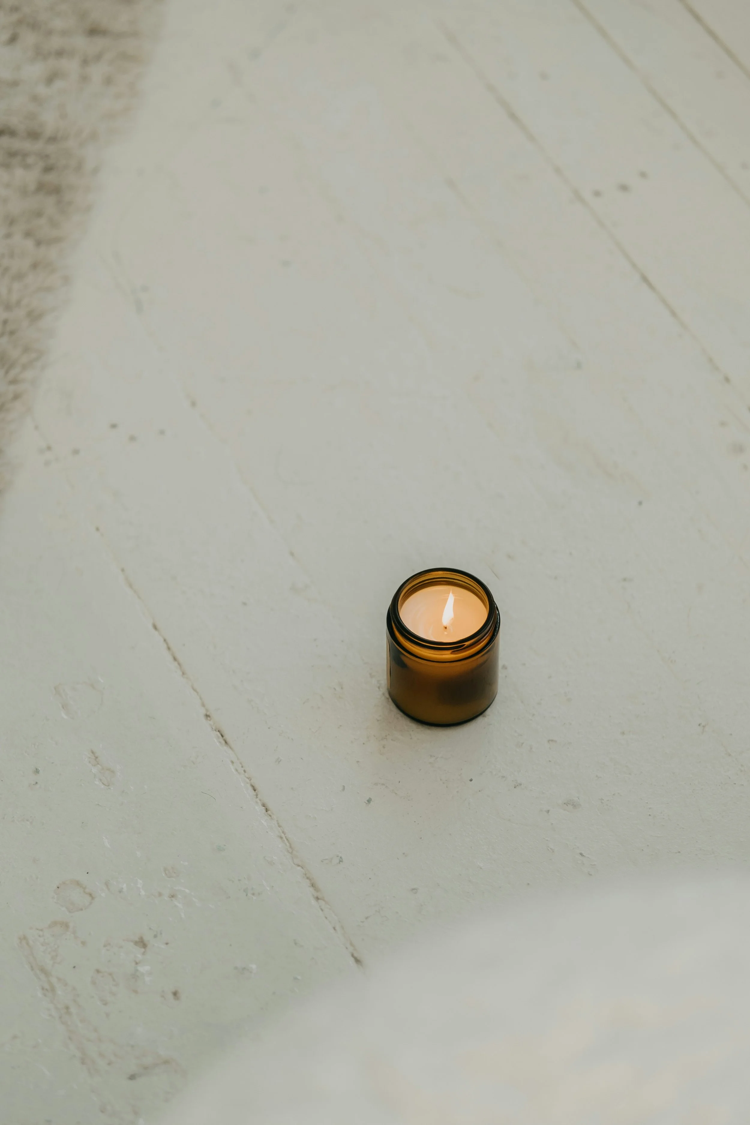 A lit candle in a small brown glass jar placed on a white wooden floor, with part of a beige rug visible in the top left corner.