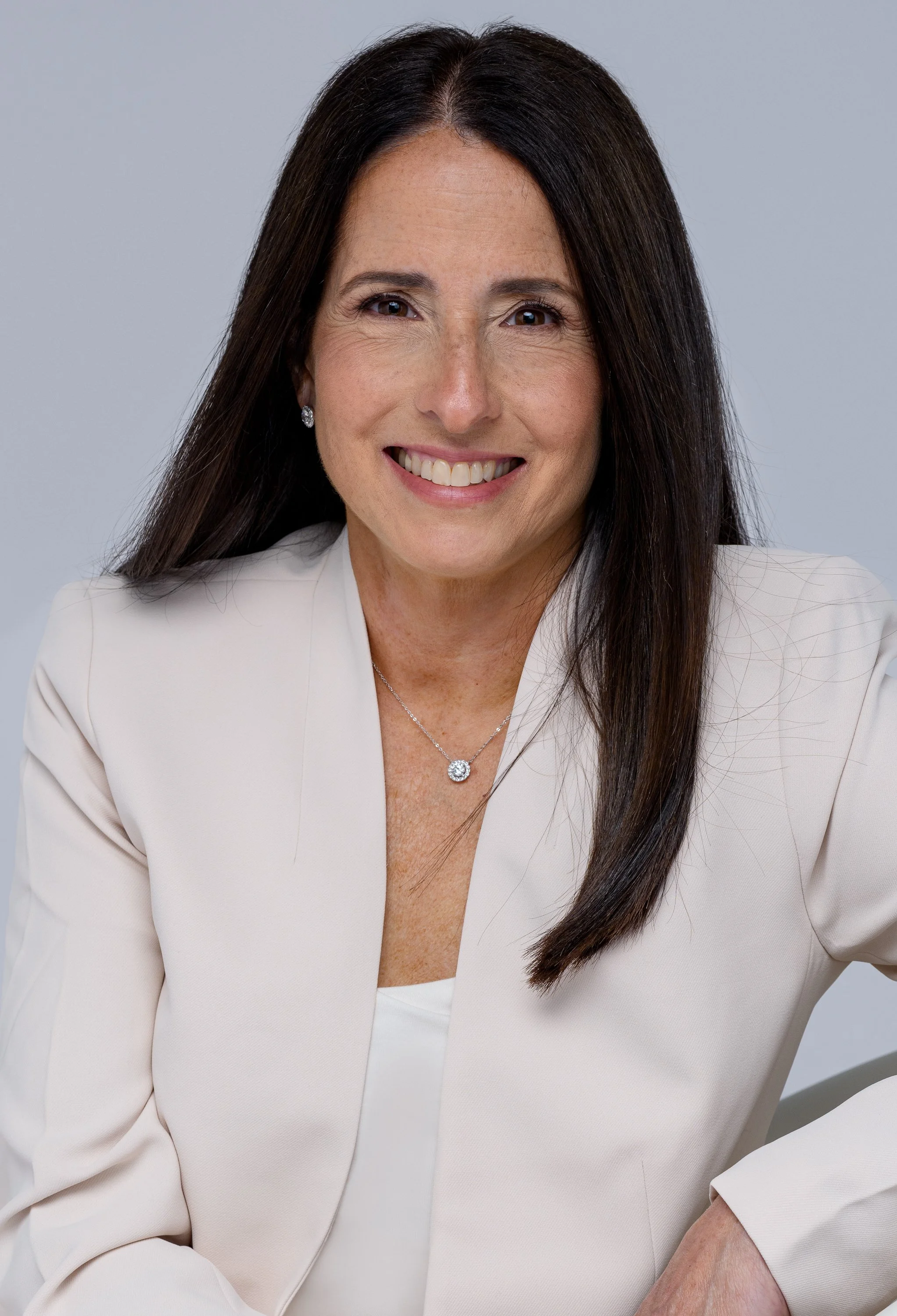 Professional woman with long dark hair, wearing a white blazer and jewelry, smiling at the camera against a neutral background.