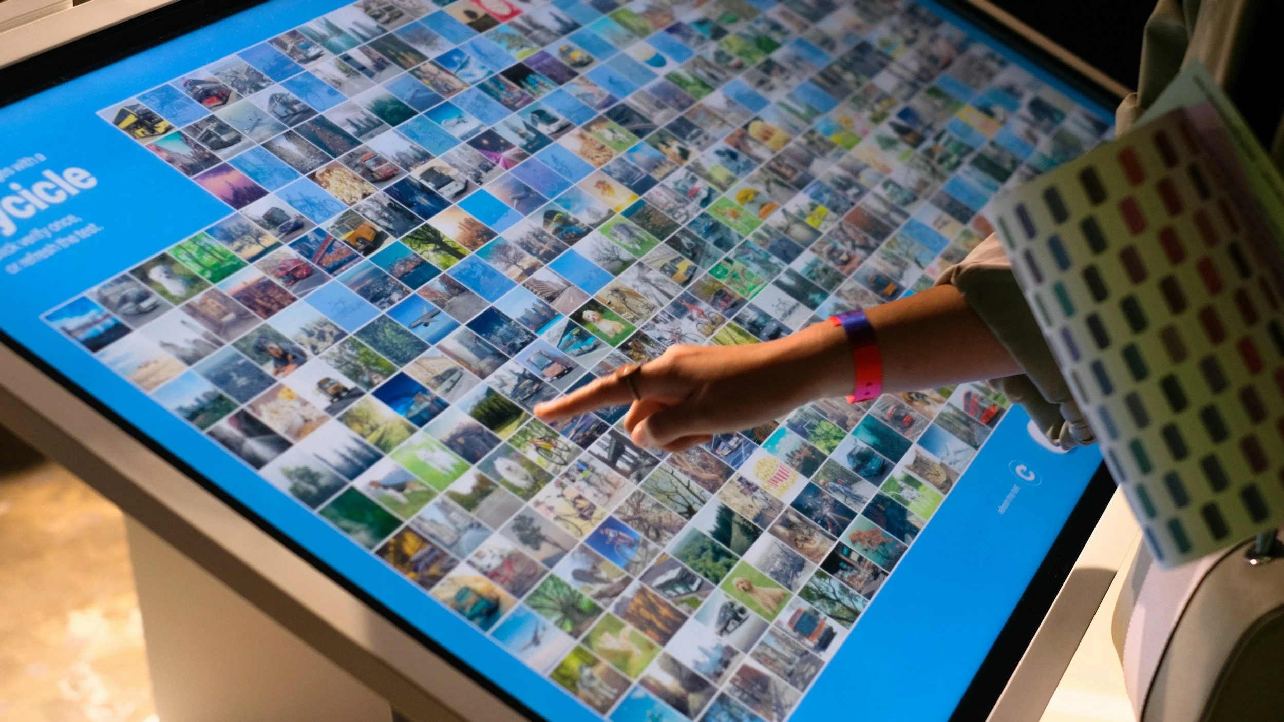 Touchscreen display with a grid of small, colorful photographs of various landscapes, cityscapes, and outdoor scenes, with a person's arm and finger interacting with it.