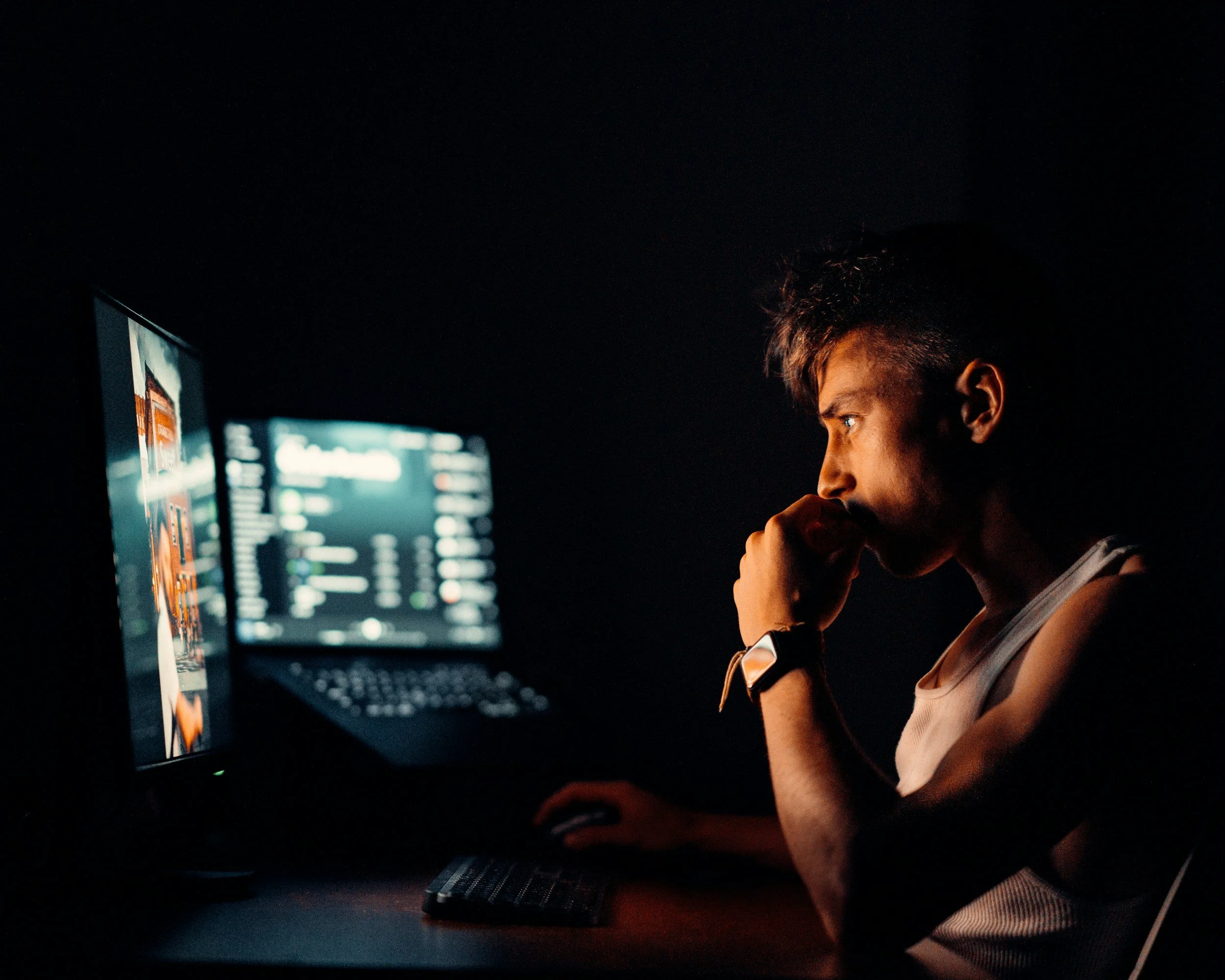 A person with short dark hair and a beard sitting at a desk in a dark room, using a computer with multiple monitors with bright screens.