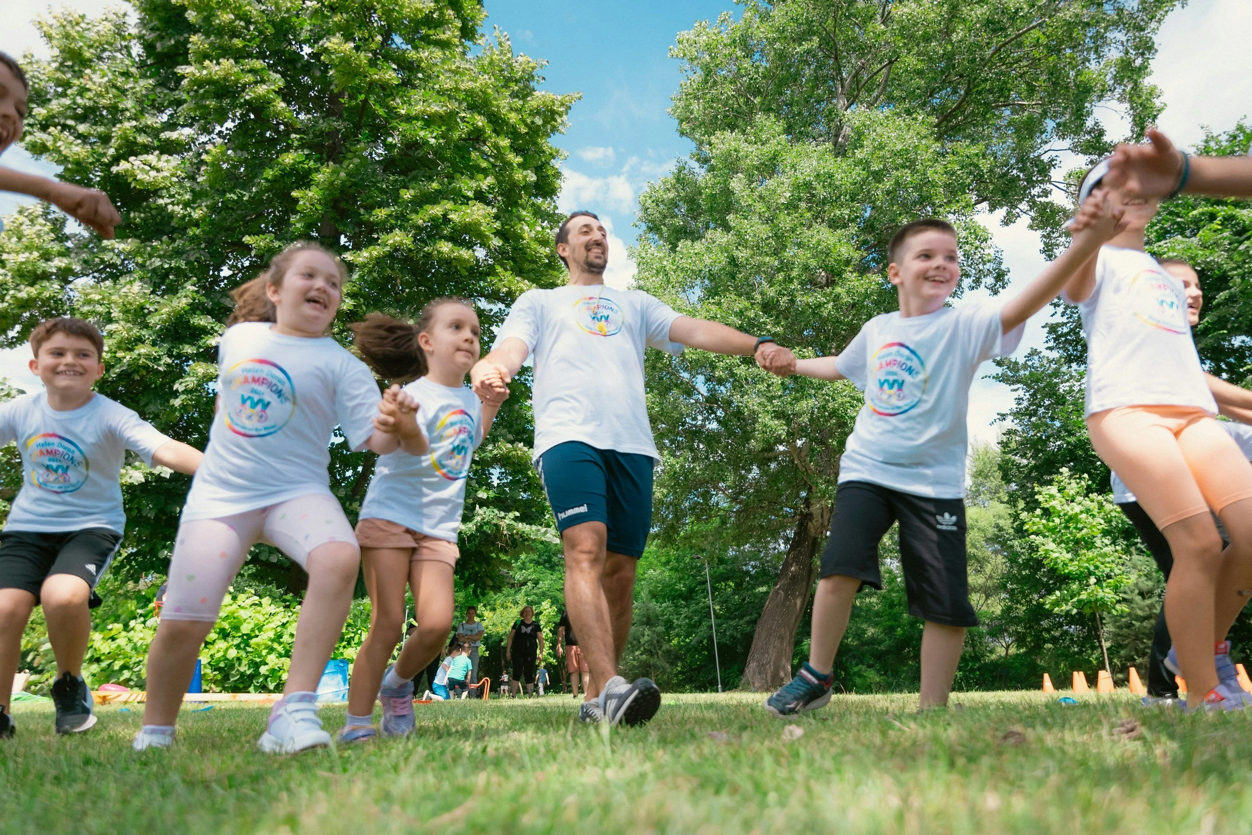 Children and an adult holding hands and jumping in a park with green trees, during a sunny day, participating in an outdoor activity or event.