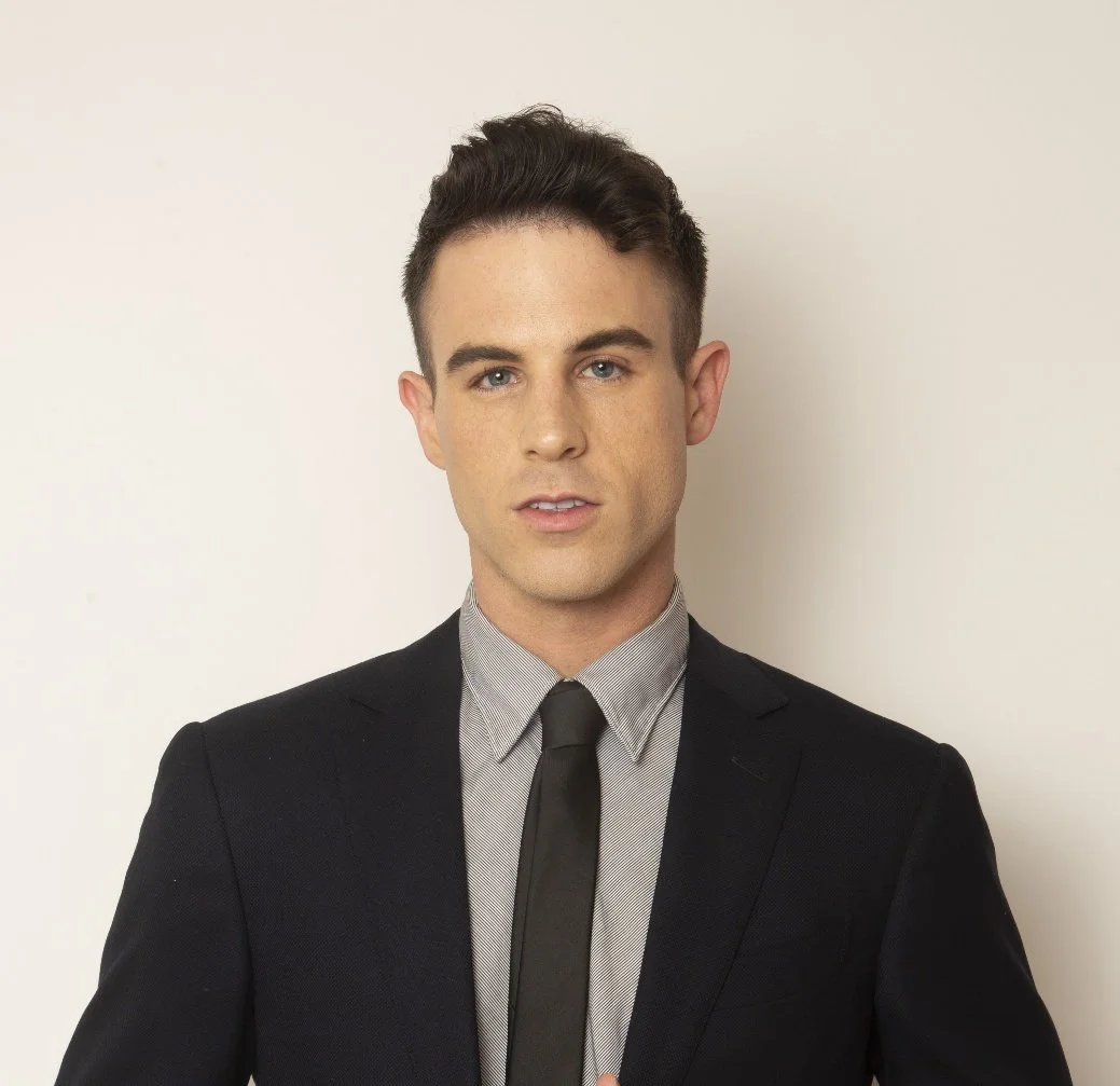 A young man wearing a black suit, light gray shirt, and black tie posing against a plain white wall.