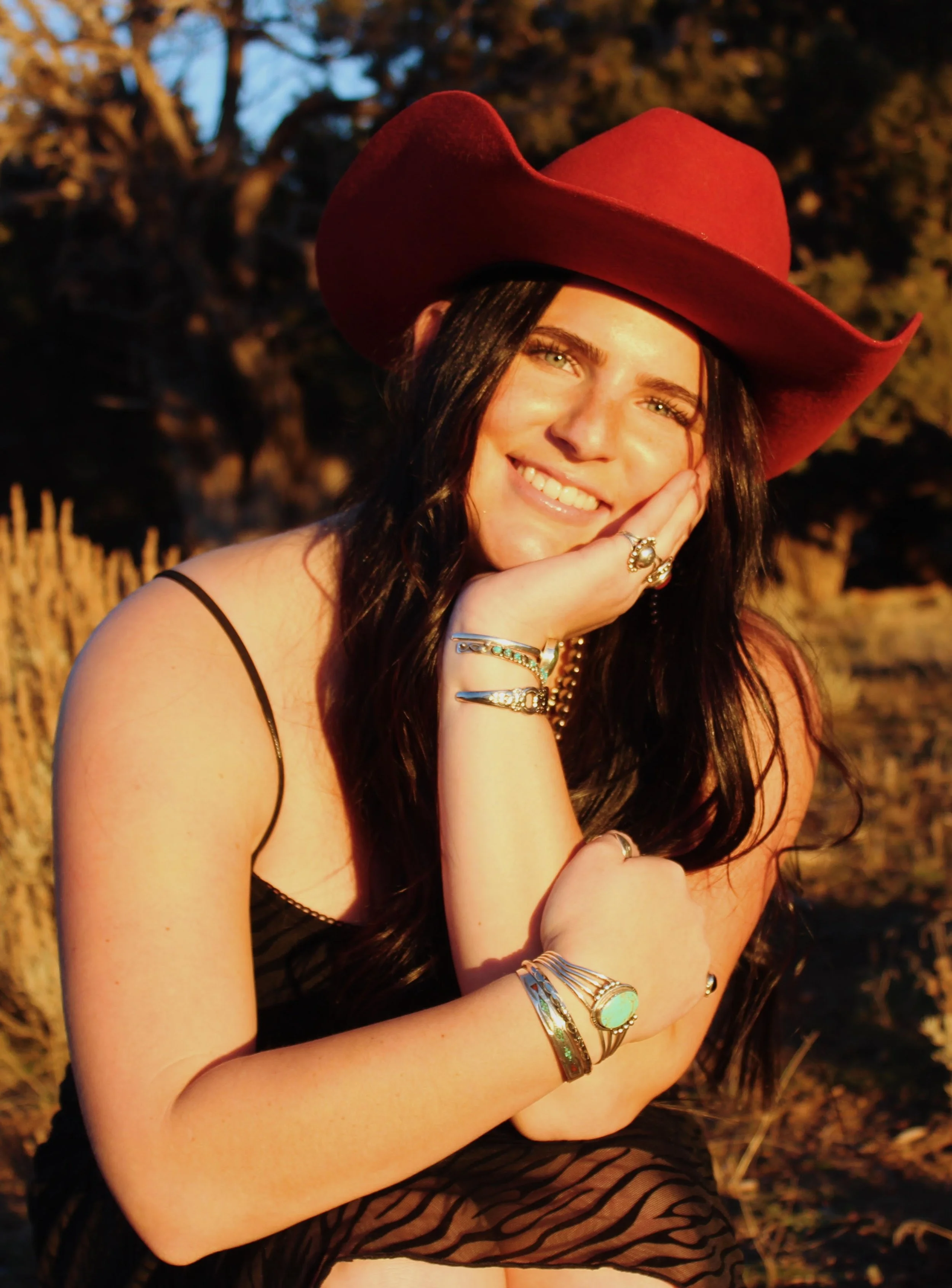 A woman with long dark hair wearing a large red cowboy hat, smiling, resting her face on her hand, outdoors during sunset, adorned with multiple rings and bracelets, wearing a black spaghetti strap dress.