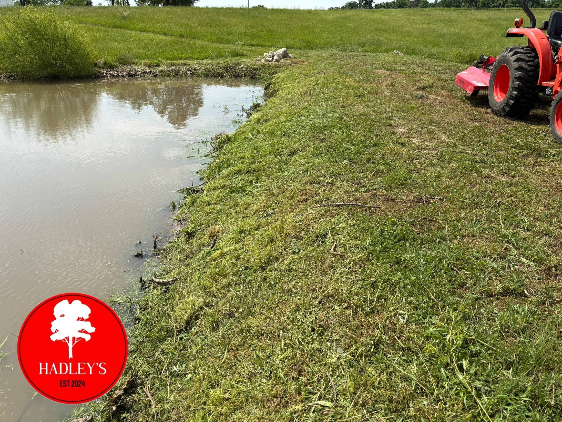 A small body of water with green grass and a dirt bank along the edge. A red tractor is parked on the grassy bank to the right, and a red and white circular sign with a tree logo and the text 'HADLEY'S EST 2024' is in the bottom left corner.