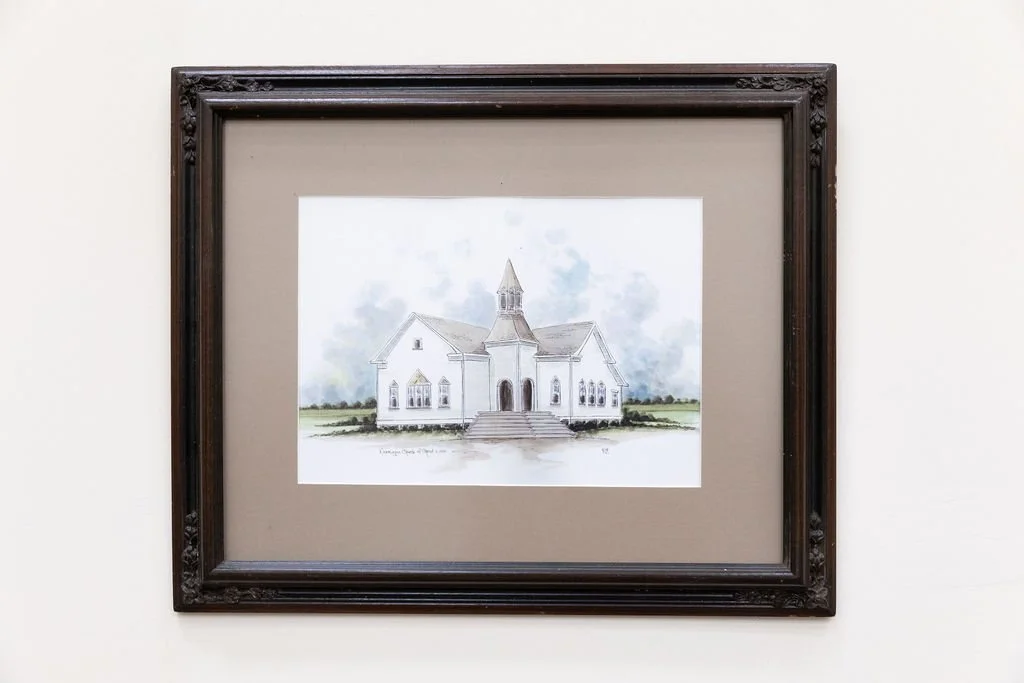 Watercolor illustration of the white Normangee church of Christ building with a steeple, steps leading to the entrance, and surrounding grassy area, framed and hung on a wall inside the building itself.