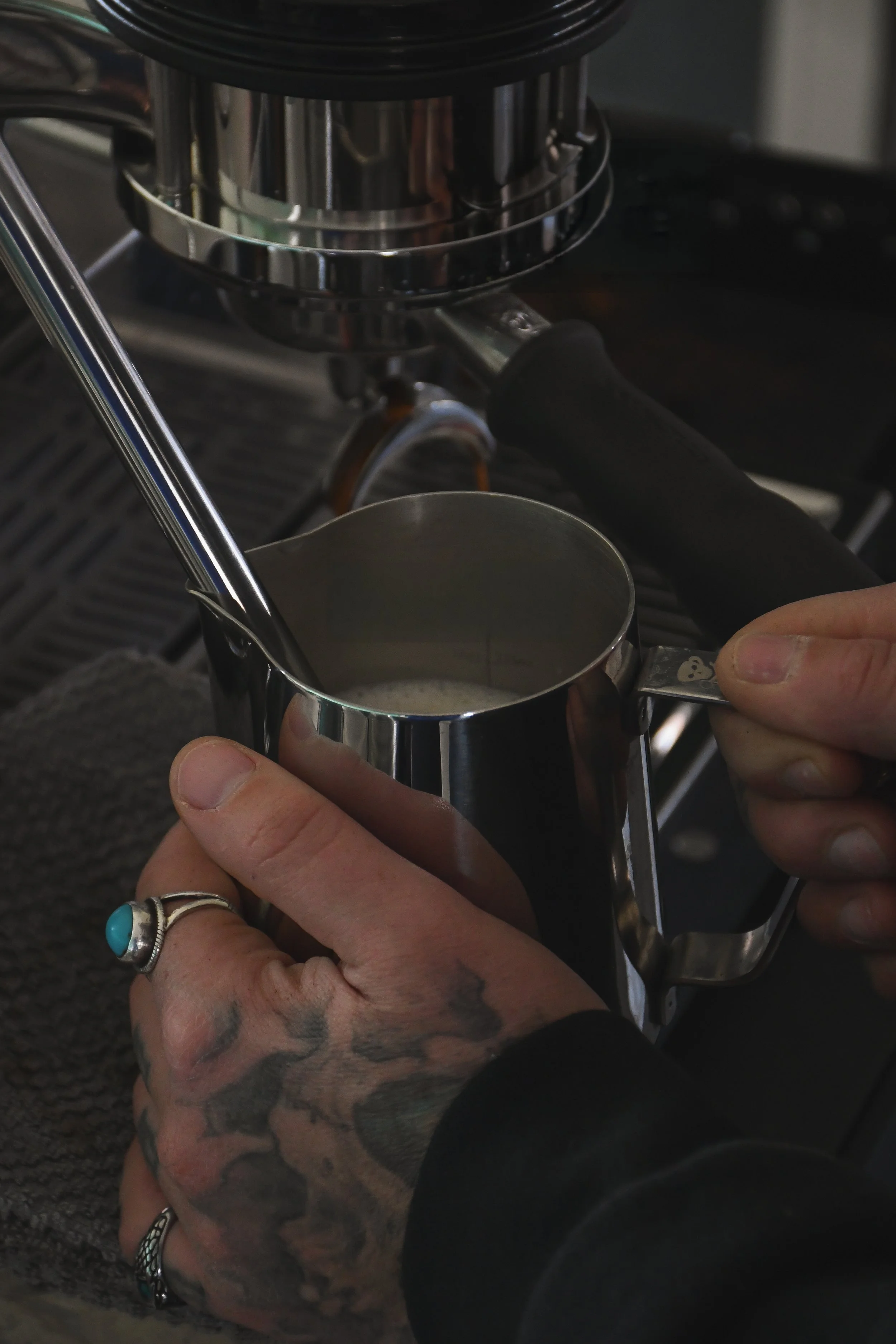 Close-up of hands with tattoos and rings holding a metal milk frothing pitcher under a coffee machine's steam wand.
