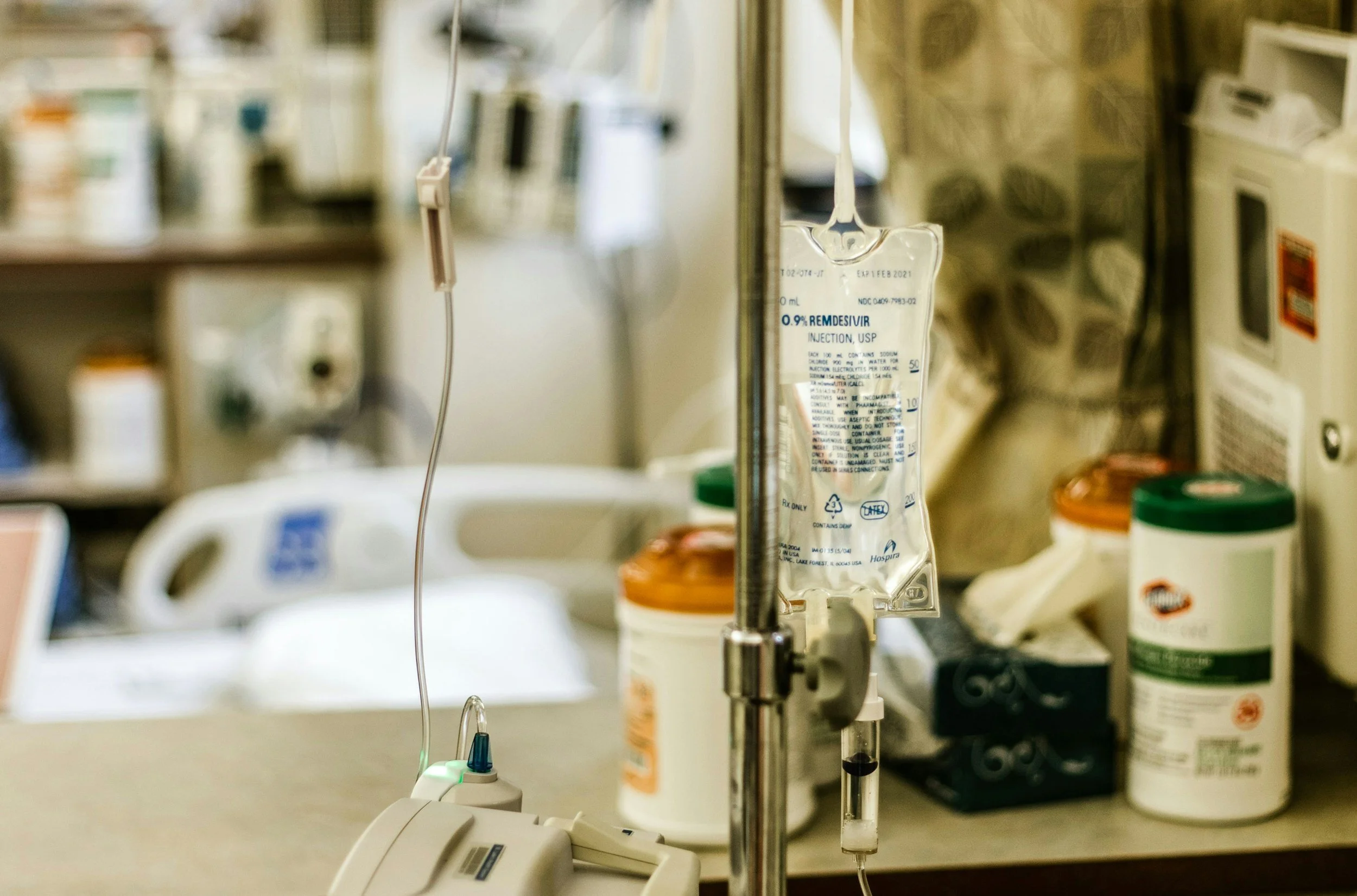 Hospital IV drip bag with medication label, medical supplies, and pharmacy bottles in the background.
