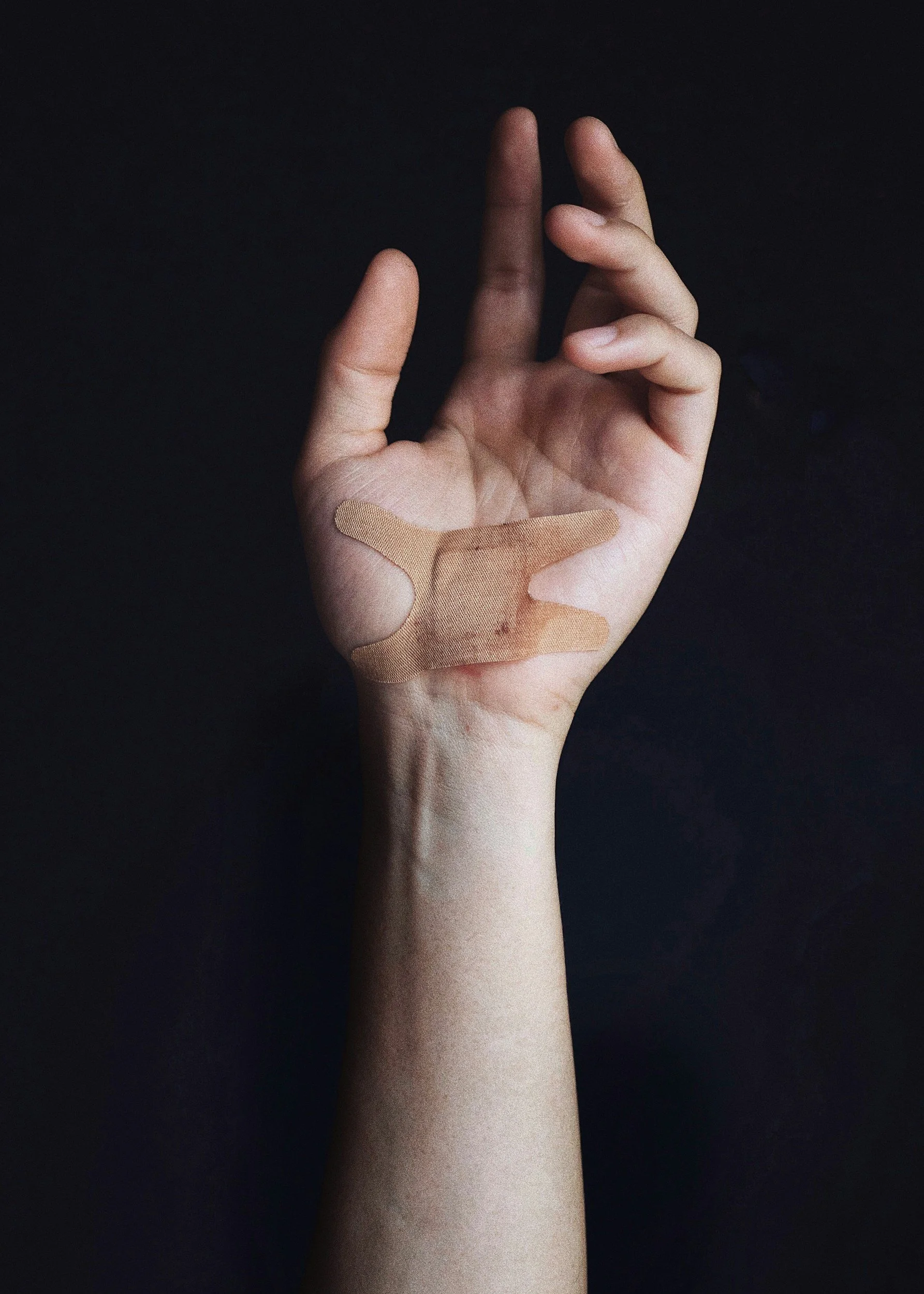 A hand with a bandage on the palm and thumb, making an 'OK' gesture against a black background.