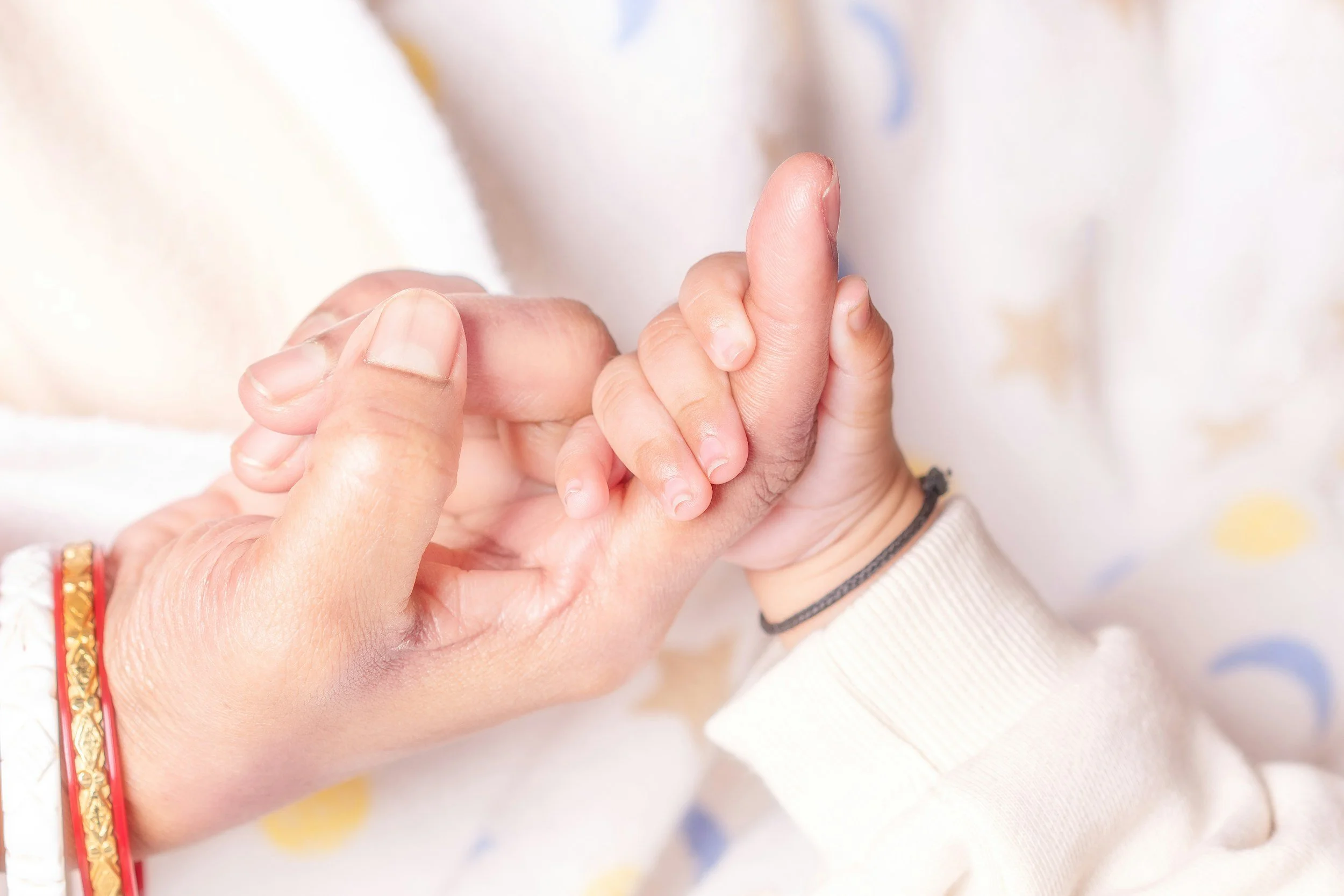 An adult hand gently holding and supporting a small child's hand, both with light skin tones. The adult's hand has a red and gold bracelet and the child's wrist has a black hair tie. The background is soft and blurred.