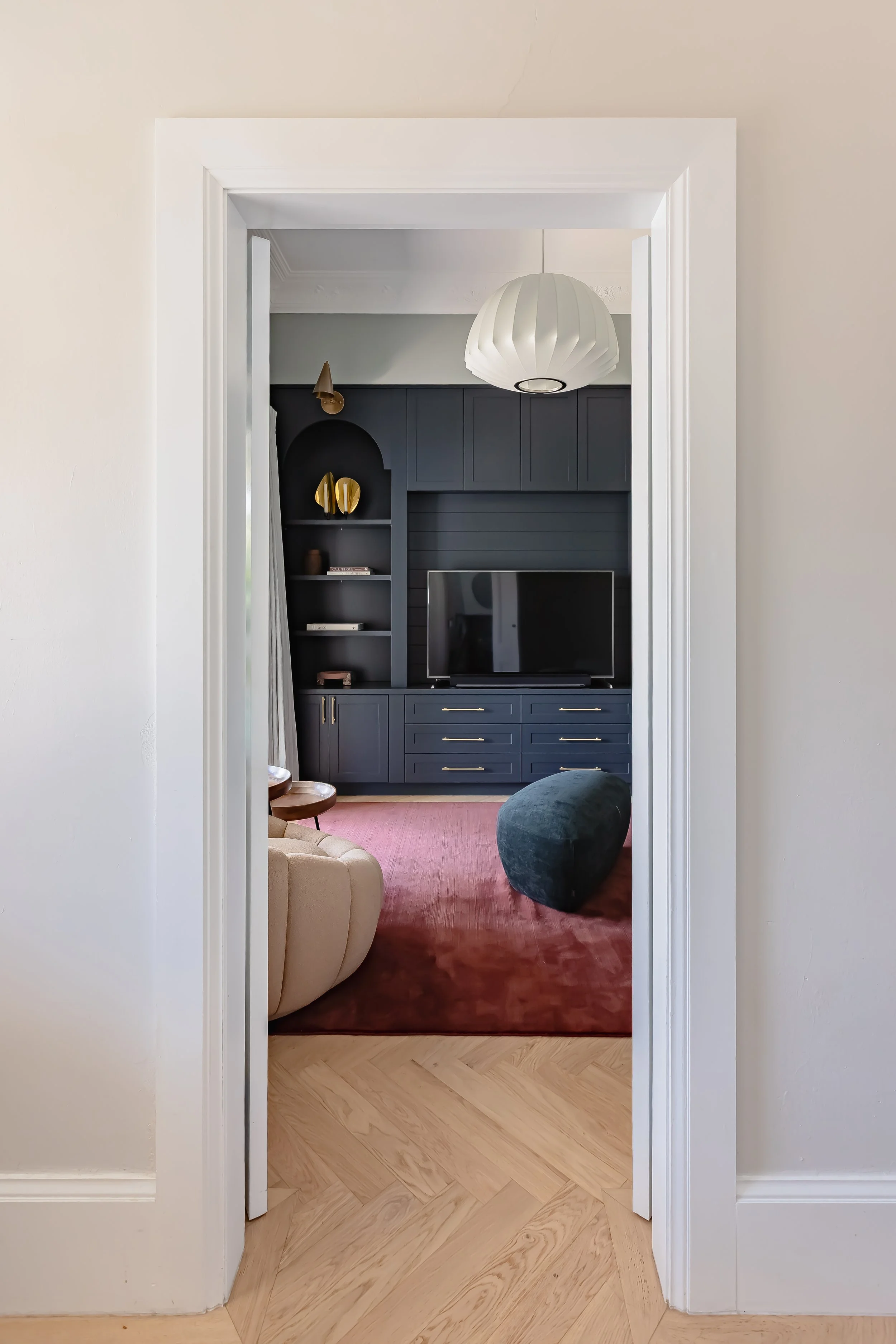 Living room with dark blue built-in shelves and cabinets, a flat-screen TV, a pink area rug, beige and dark green seating, and a white paper lantern ceiling light.