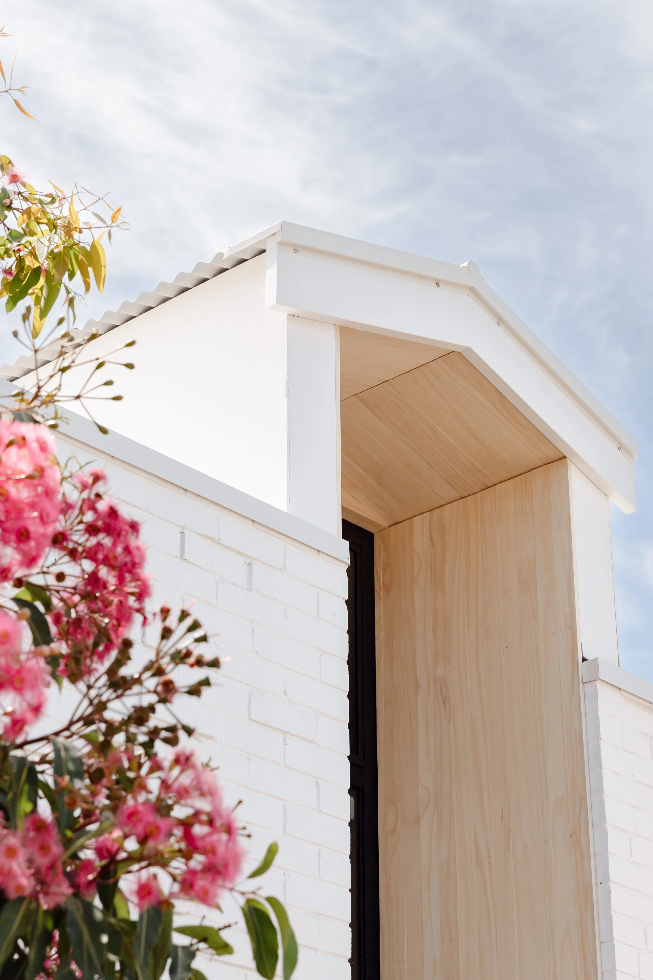 Close-up of a modern building with white brick walls, wooden accents, and a roof, with pink flowering plants in the foreground and a sky with scattered clouds in the background.